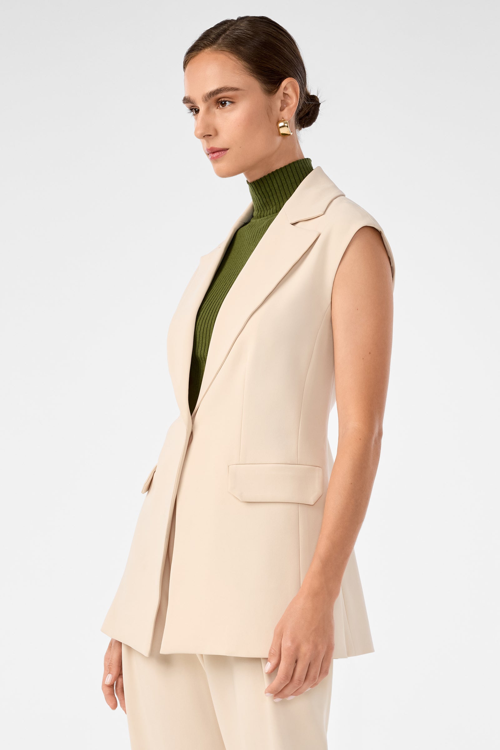 A woman stands against a light background, wearing the Boabab Sleeveless Jacket in beige over a green turtleneck and matching beige trousers. Her hair is pulled back as she looks to the side.