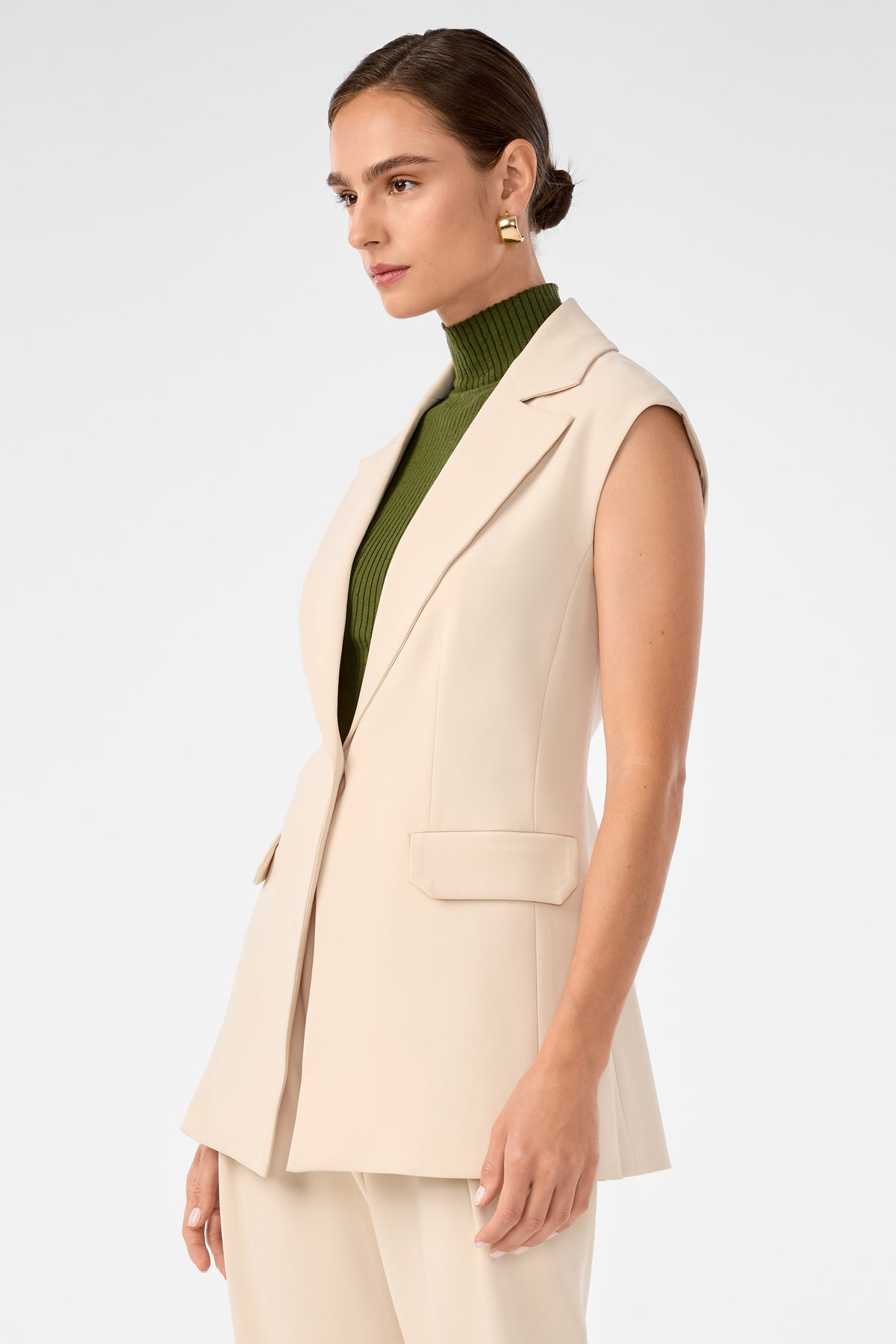 A woman stands against a light background, wearing the Boabab Sleeveless Jacket in beige over a green turtleneck and matching beige trousers. Her hair is pulled back as she looks to the side.