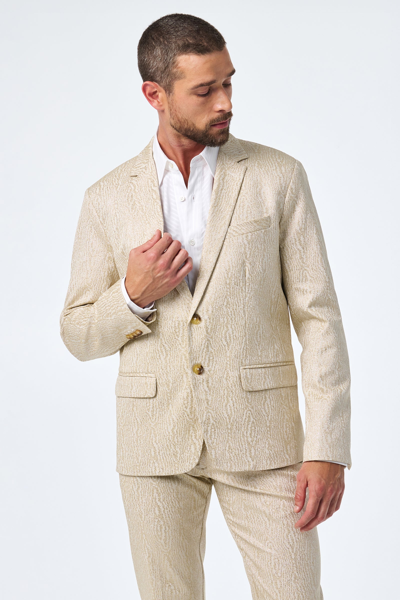 A man with short hair and a trimmed beard wears the light beige, textured Thurston Blazer over a white dress shirt. He looks down left, adjusting his jacket against a plain white background.