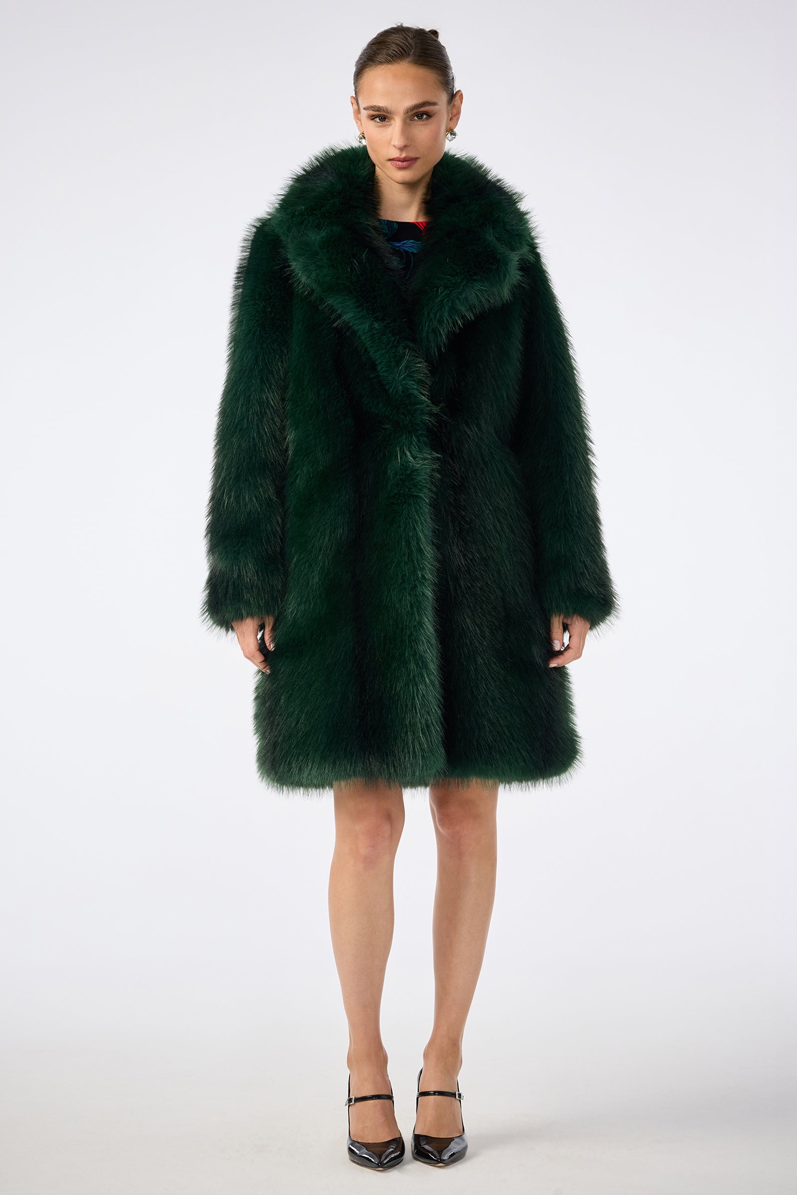 A woman faces forward in the Dayos Coat, a luxurious dark green faux fur design with a wide collar and long sleeves. She pairs it with black pointed-toe heels against a plain white background.