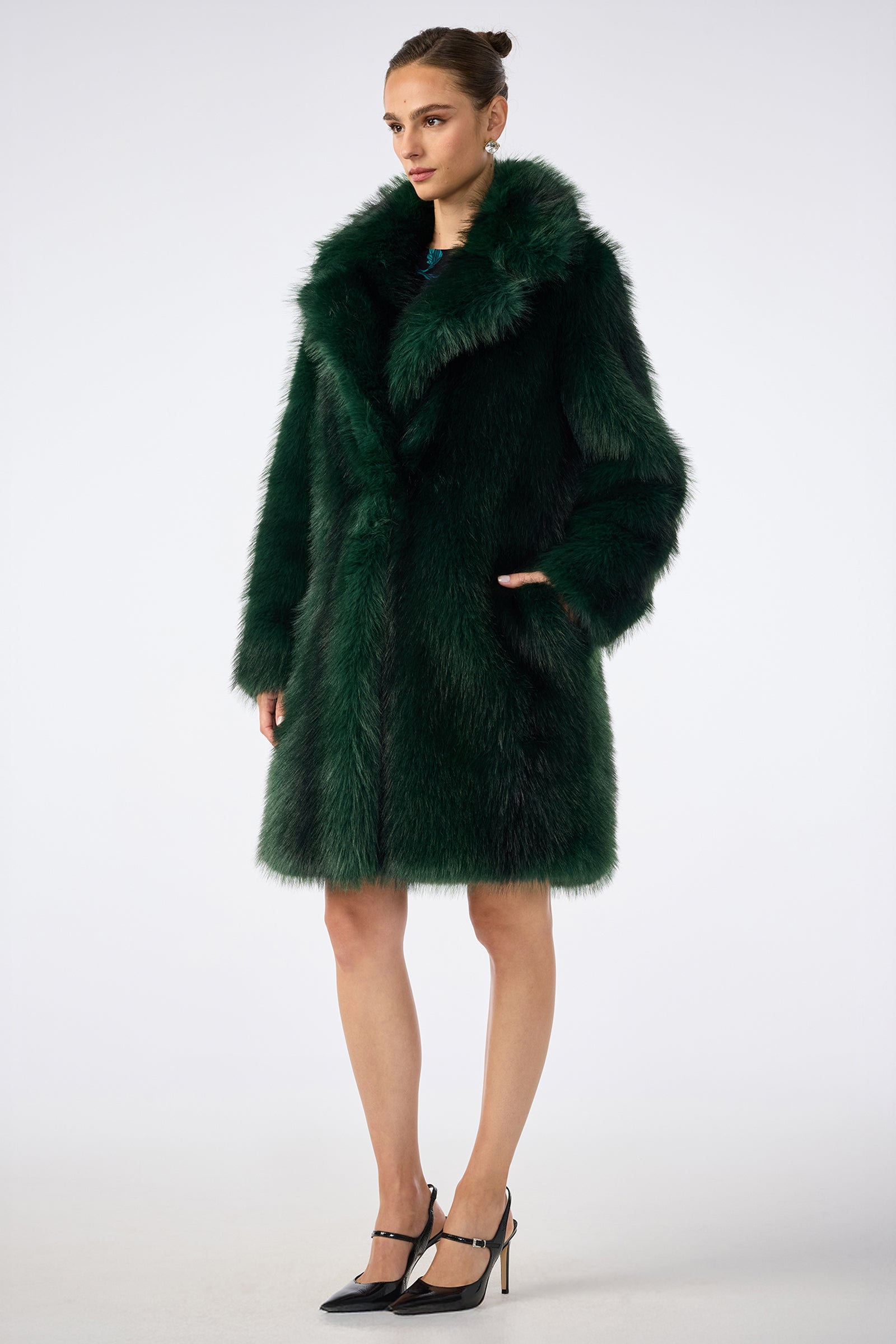 A woman with her hair in a bun stands against a plain background, wearing the Dayos Coat in dark green faux fur and black high heels. She has one hand in her pocket and a neutral expression.