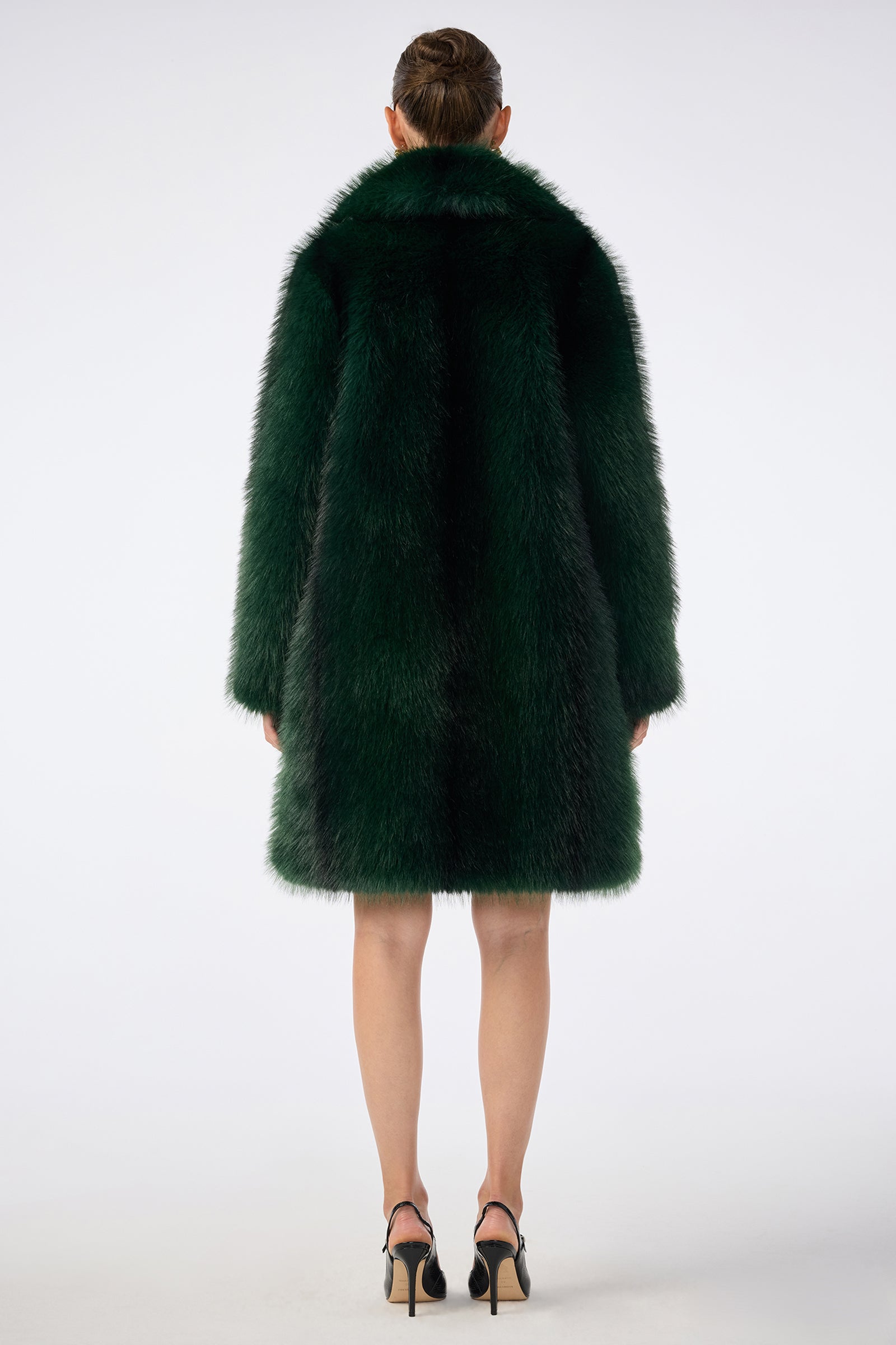A person with their hair in a bun stands facing away, wearing the long dark green Dayos Coat and black high-heeled shoes against a plain white background.