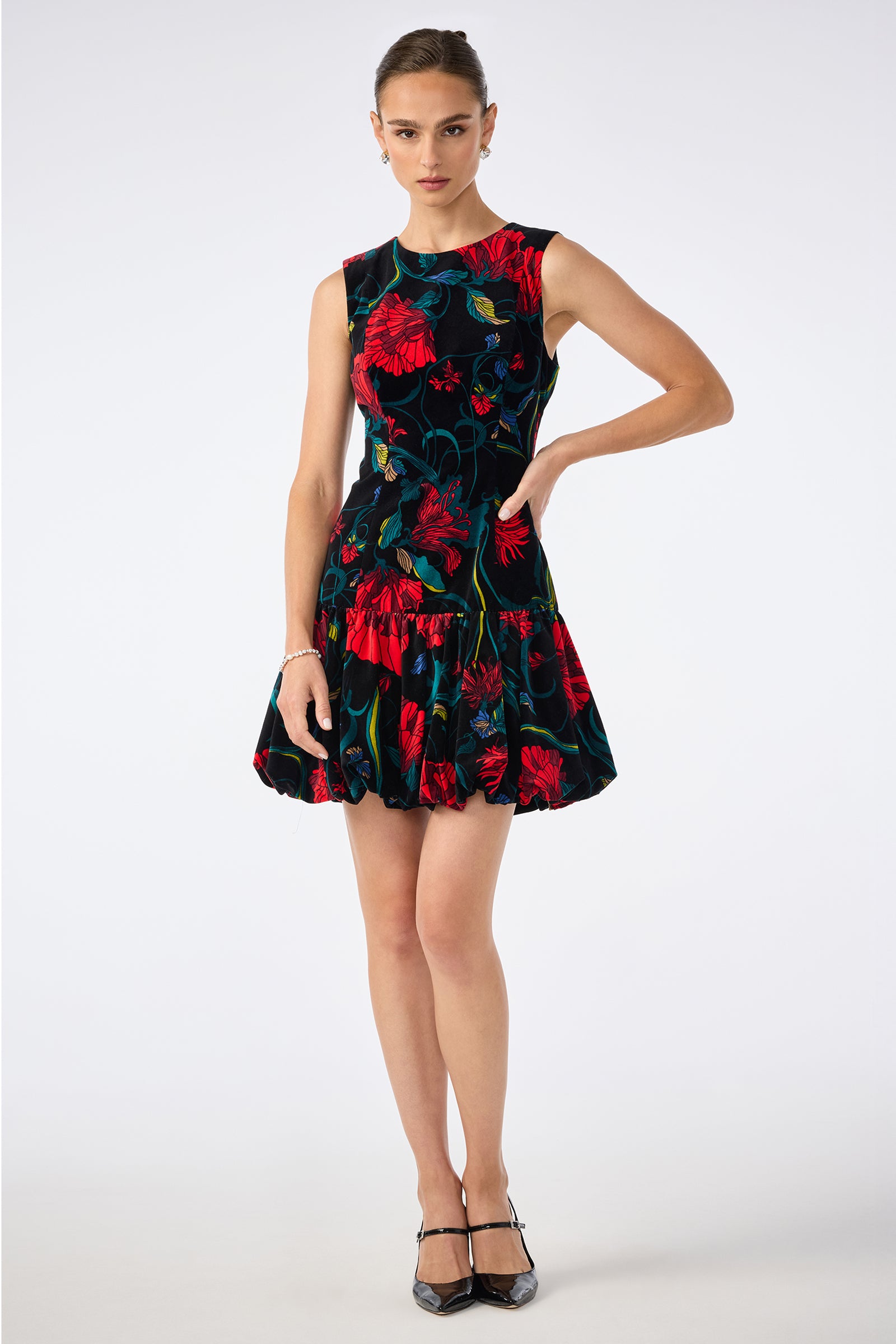 A woman poses against a plain white background in the Splendored Dress, a sleeveless black dress with a flared skirt and bold red and green floral pattern, paired with black heels and her hair tied back.
