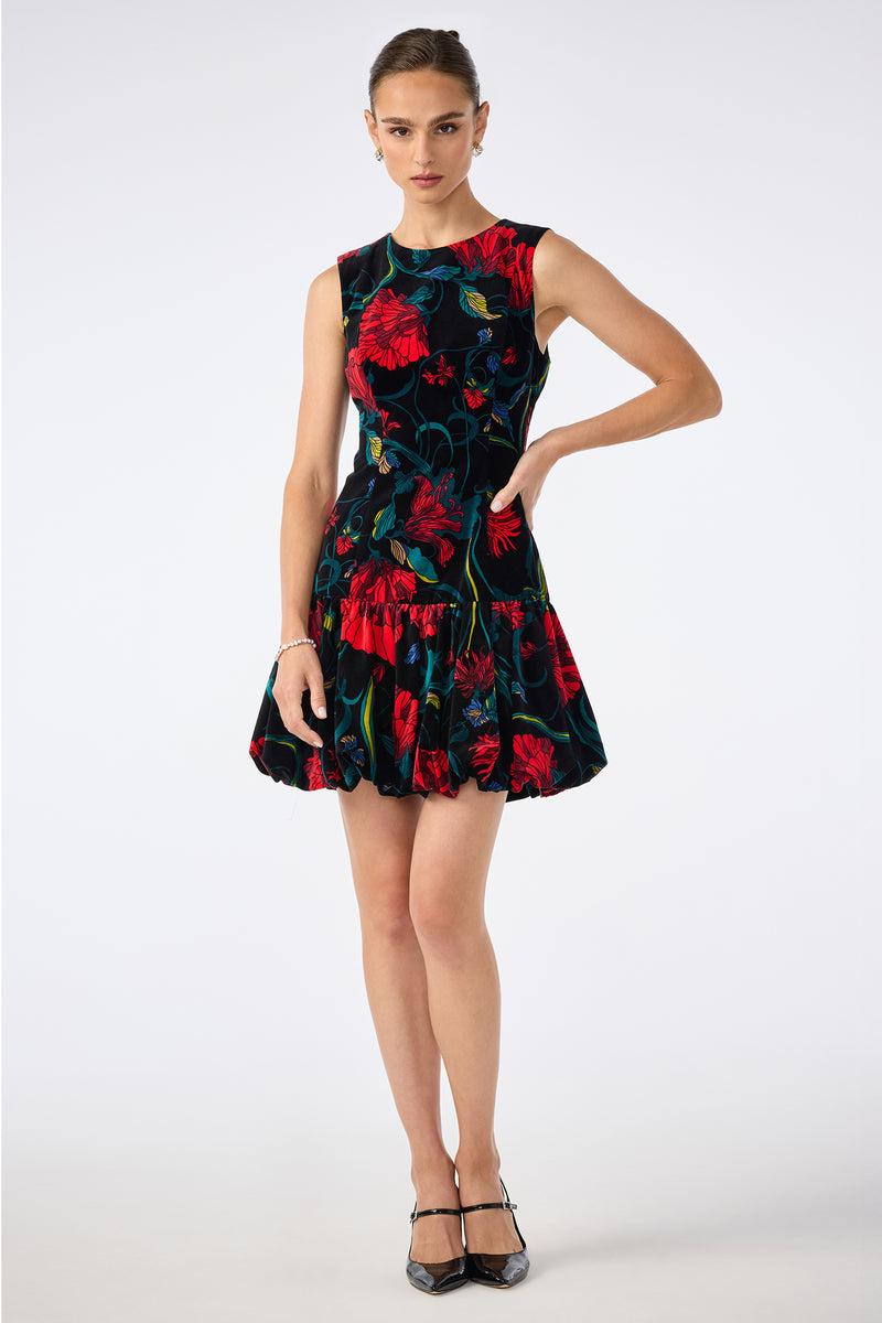 A woman poses against a plain white background in the Splendored Dress, a sleeveless black dress with a flared skirt and bold red and green floral pattern, paired with black heels and her hair tied back.
