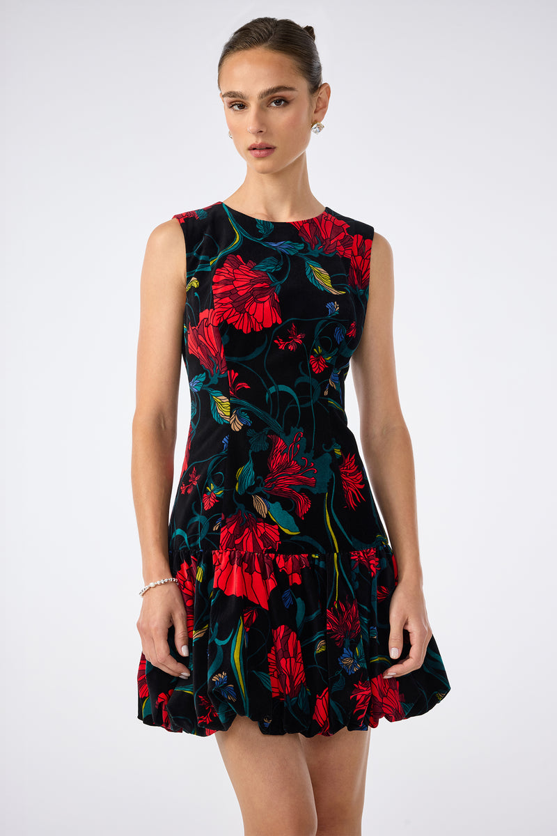 A woman wearing the Splendored Dress, a sleeveless black dress adorned with large red and blue floral patterns, stands against a plain light background.