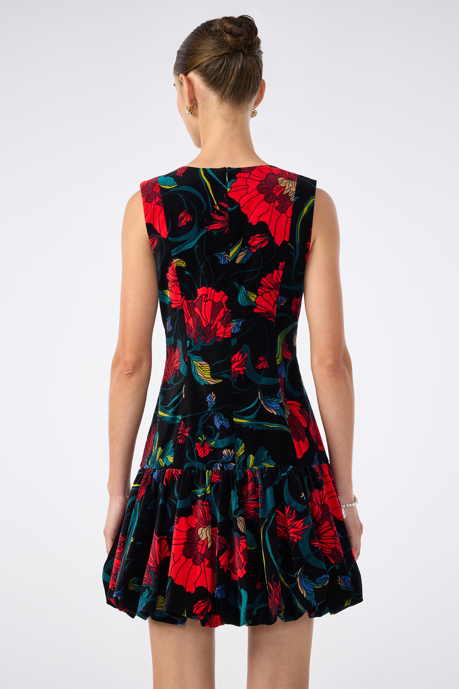 A woman with her hair in a bun models the Splendored Dress—a sleeveless black dress with a colorful floral pattern and ruffled skirt—shown from the back against a plain light background.