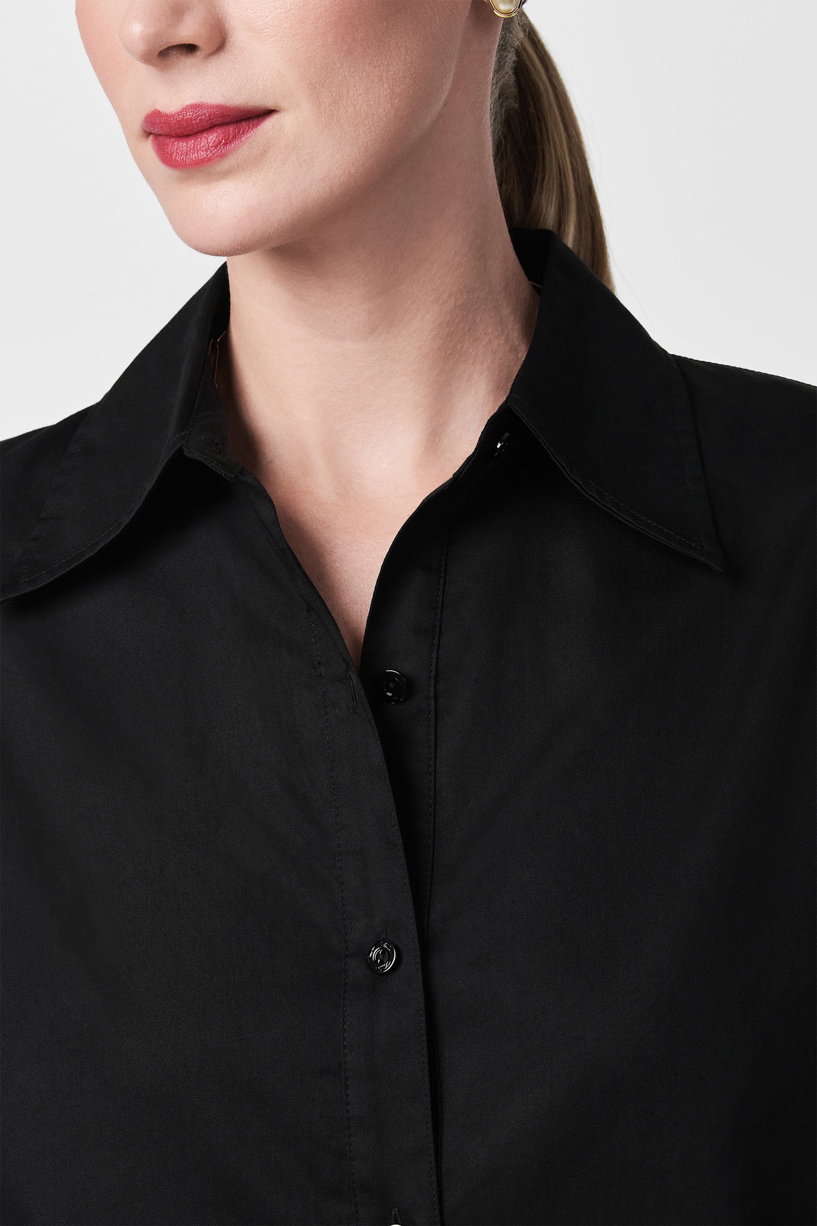 Cropped image of a woman wearing the Litsa Button-Up Cotton Shirt in black. She has light skin, pink lipstick, and brown hair pulled back; only her lower face and upper torso are visible.