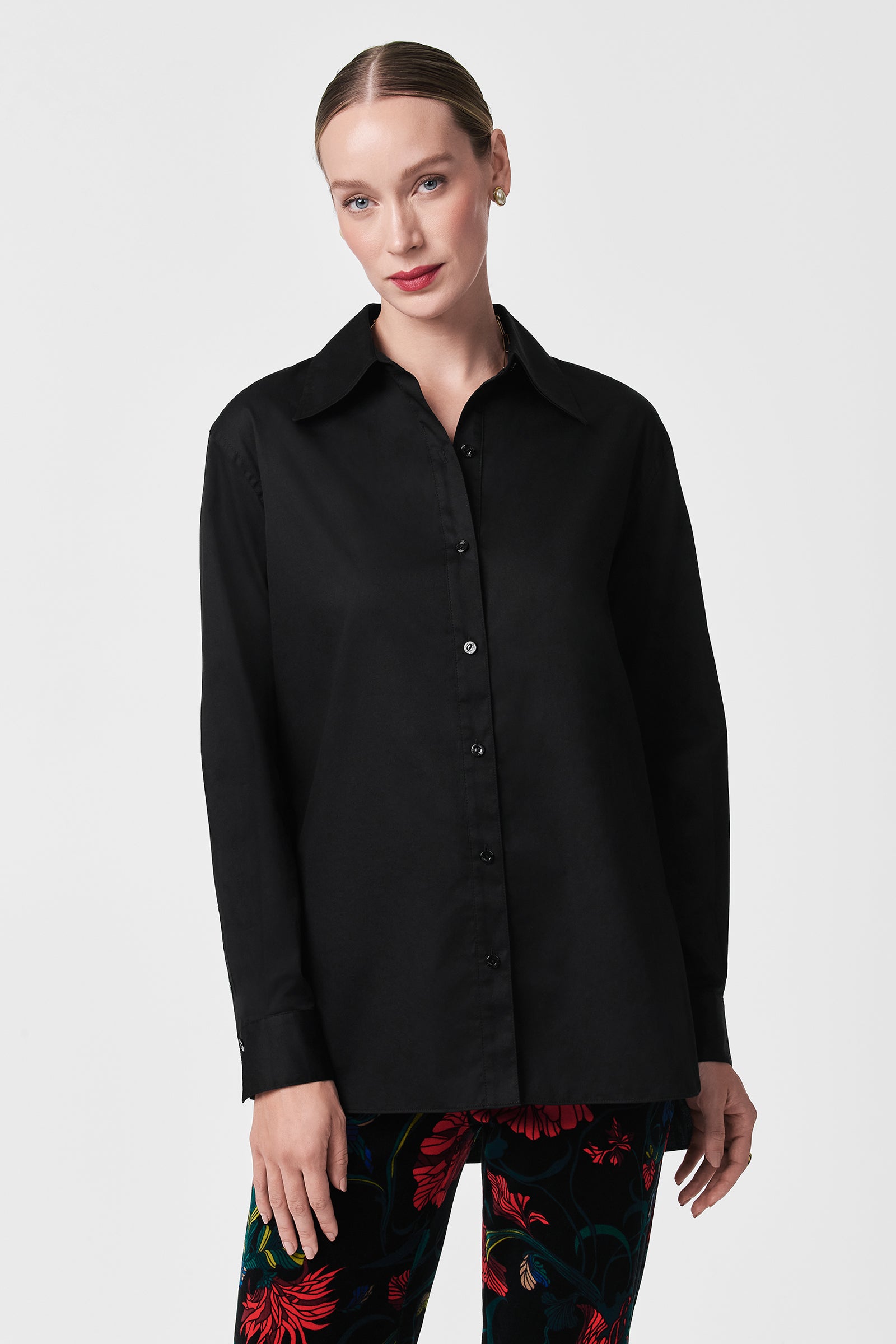 A woman with light skin and blond hair tied back wears the Litsa Button-Up Cotton Shirt in black, paired with black pants featuring a red floral pattern, standing against a plain white background.