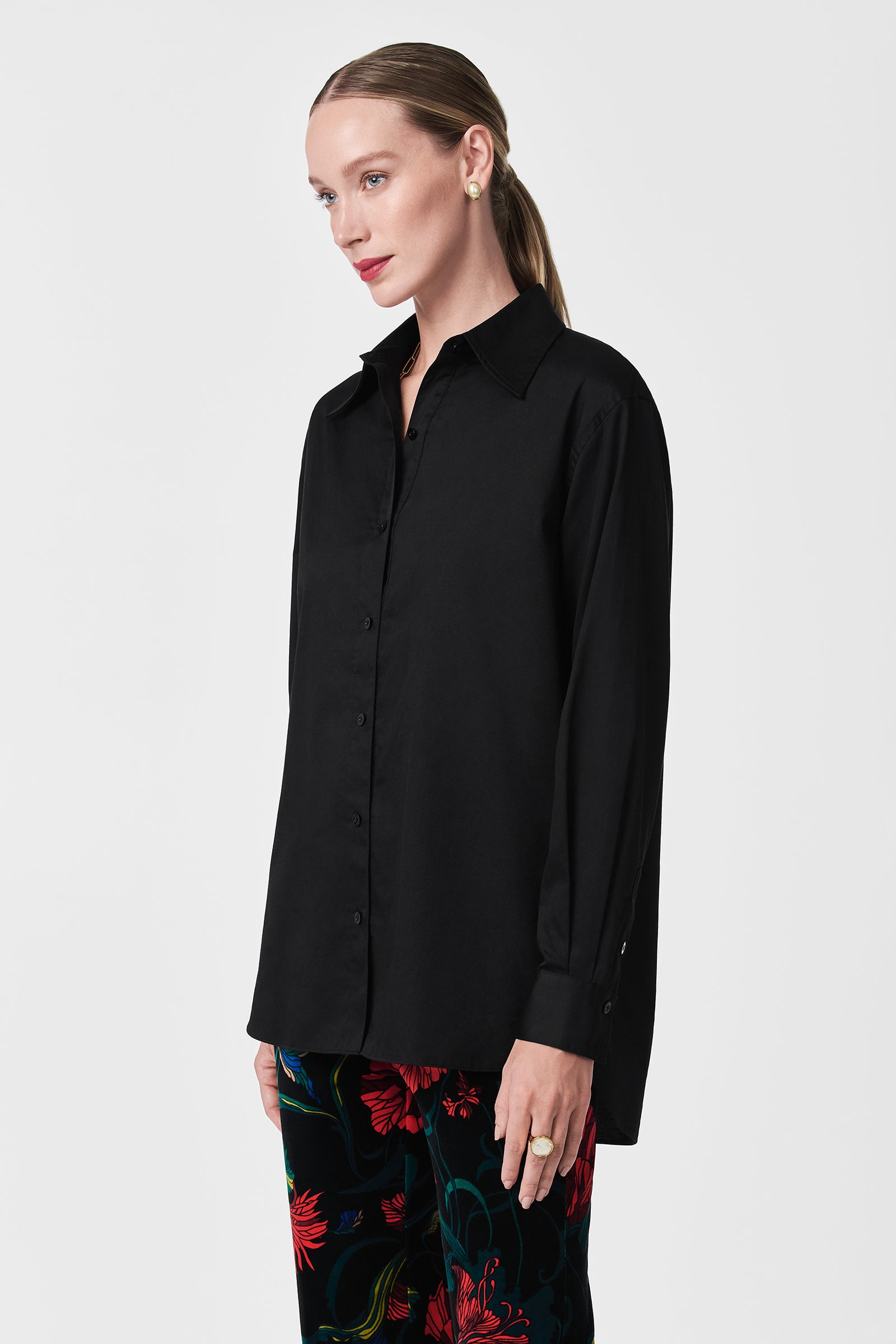 A woman with light skin and long, straight, light brown hair wears the Litsa Button-Up Cotton Shirt in black and colorful floral pants, posing against a plain white background.