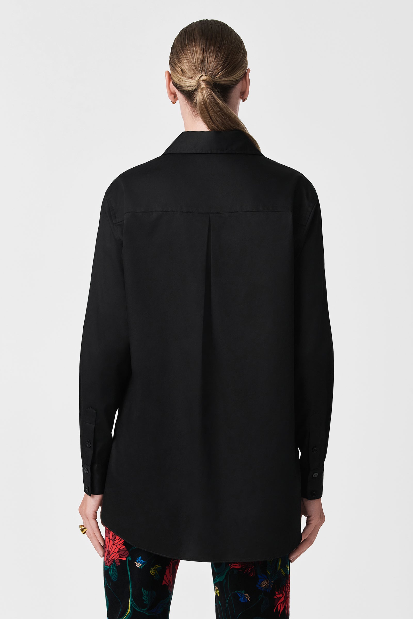 A person with long, light brown hair tied back is seen from behind wearing the loose-fitting Litsa Button-Up Cotton Shirt in black and vibrant floral pants, set against a plain white background.