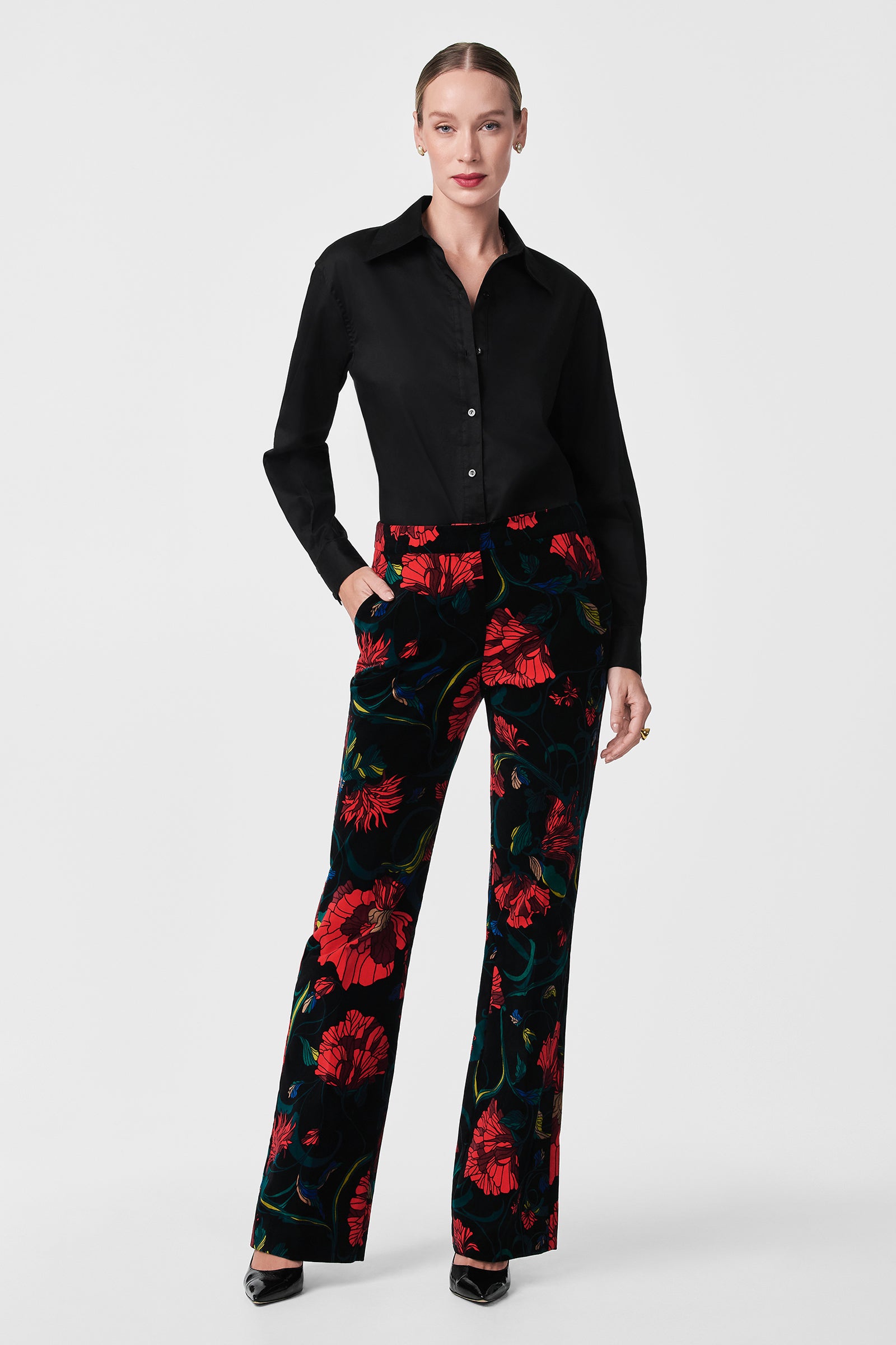 A woman stands against a plain background, wearing the Litsa Button-Up Cotton Shirt paired with black pants featuring a bold red floral print. Her hands are in her pockets and she completes the look with black heels.