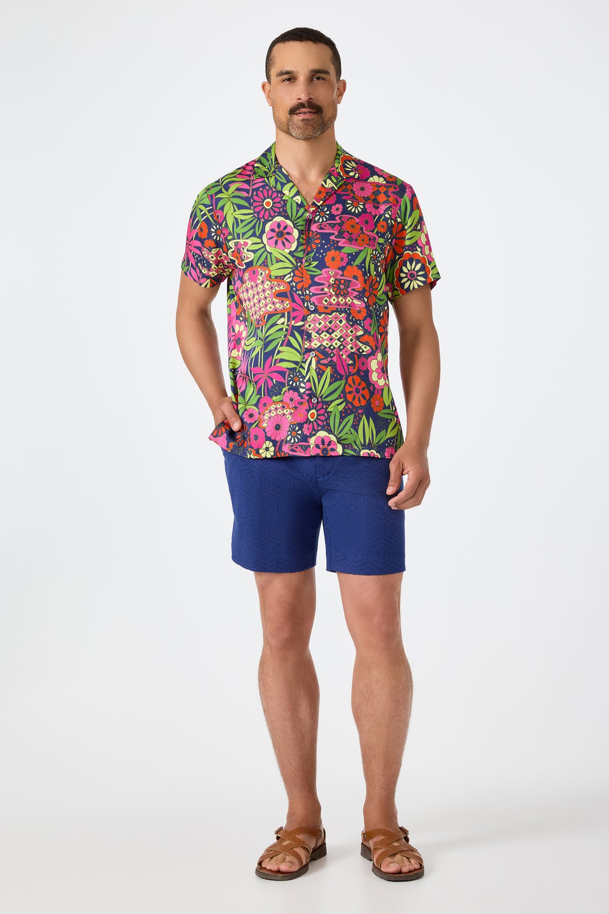 A man with short hair and a mustache stands against a white background wearing the Lawrence Short - Noh Nightfall, a colorful floral short-sleeve shirt, blue shorts, and brown sandals, with one hand in his pocket.
