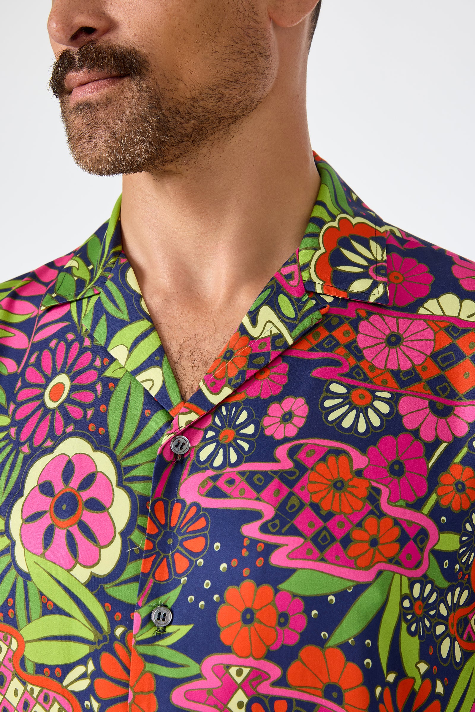 A man with a mustache wears the Felix Camp Shirt - Fumi Floral, featuring a vibrant floral pattern in purple, pink, red, orange, and green. The image is cropped to show only the lower half of his face and upper torso.