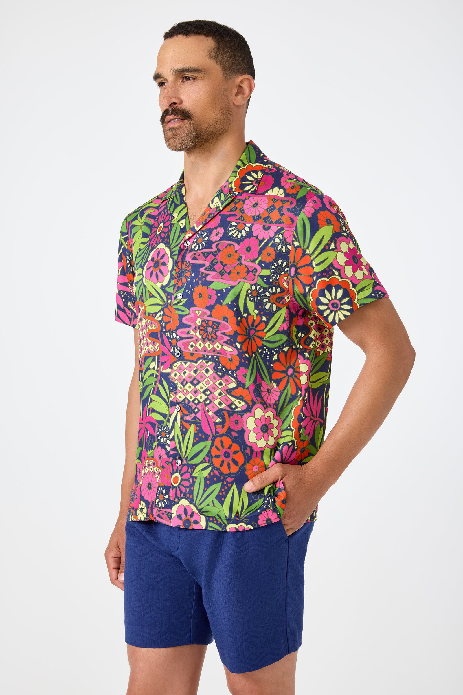 A man with short hair and a mustache wears the Felix Camp Shirt - Fumi Floral, featuring colorful green, pink, orange, and purple patterns with blue shorts, standing against a plain white background.