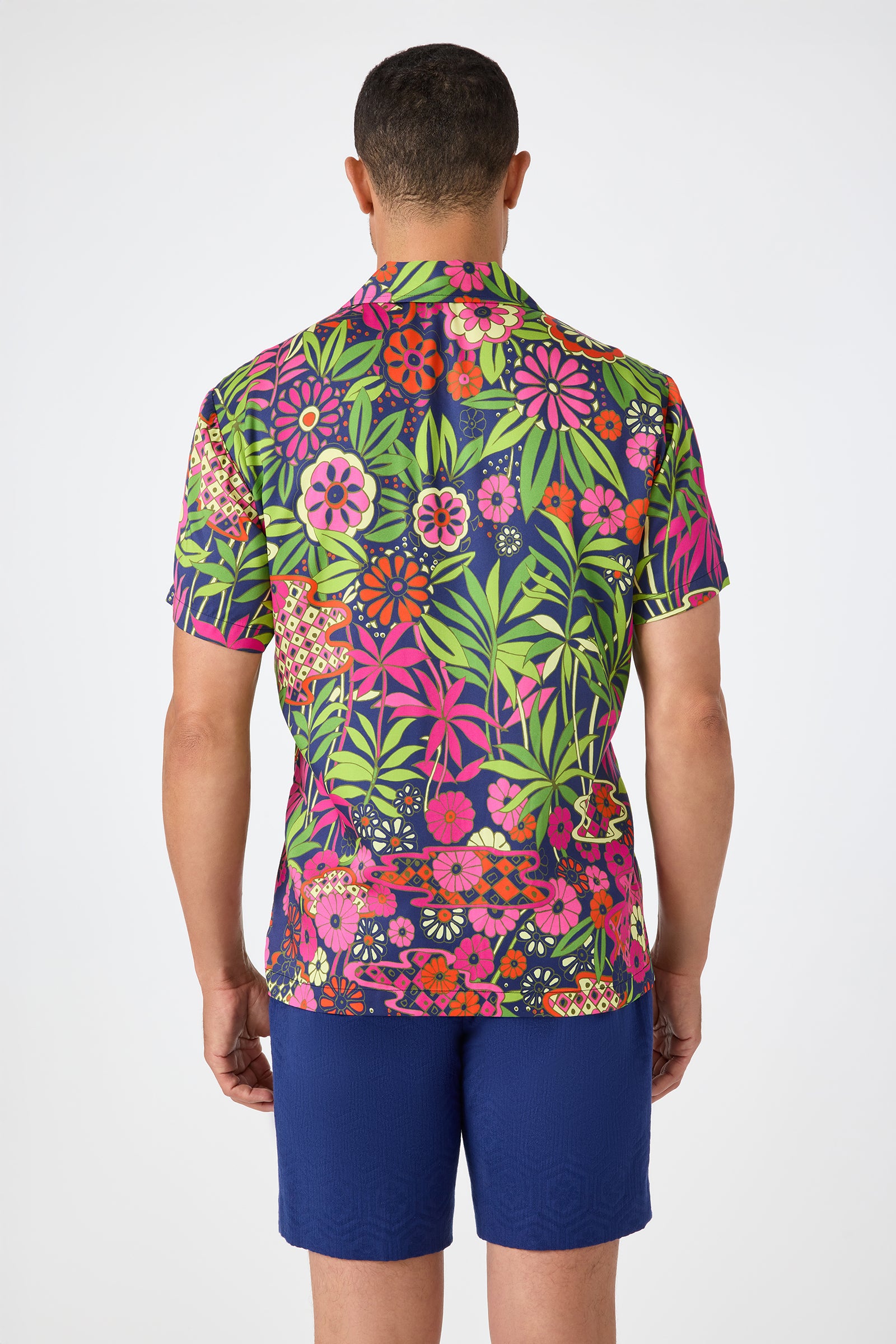 A man stands facing away, wearing the Felix Camp Shirt - Fumi Floral, a vibrant short-sleeve shirt with bold pink, green, and orange floral and leaf patterns, paired with solid blue shorts against a white background.