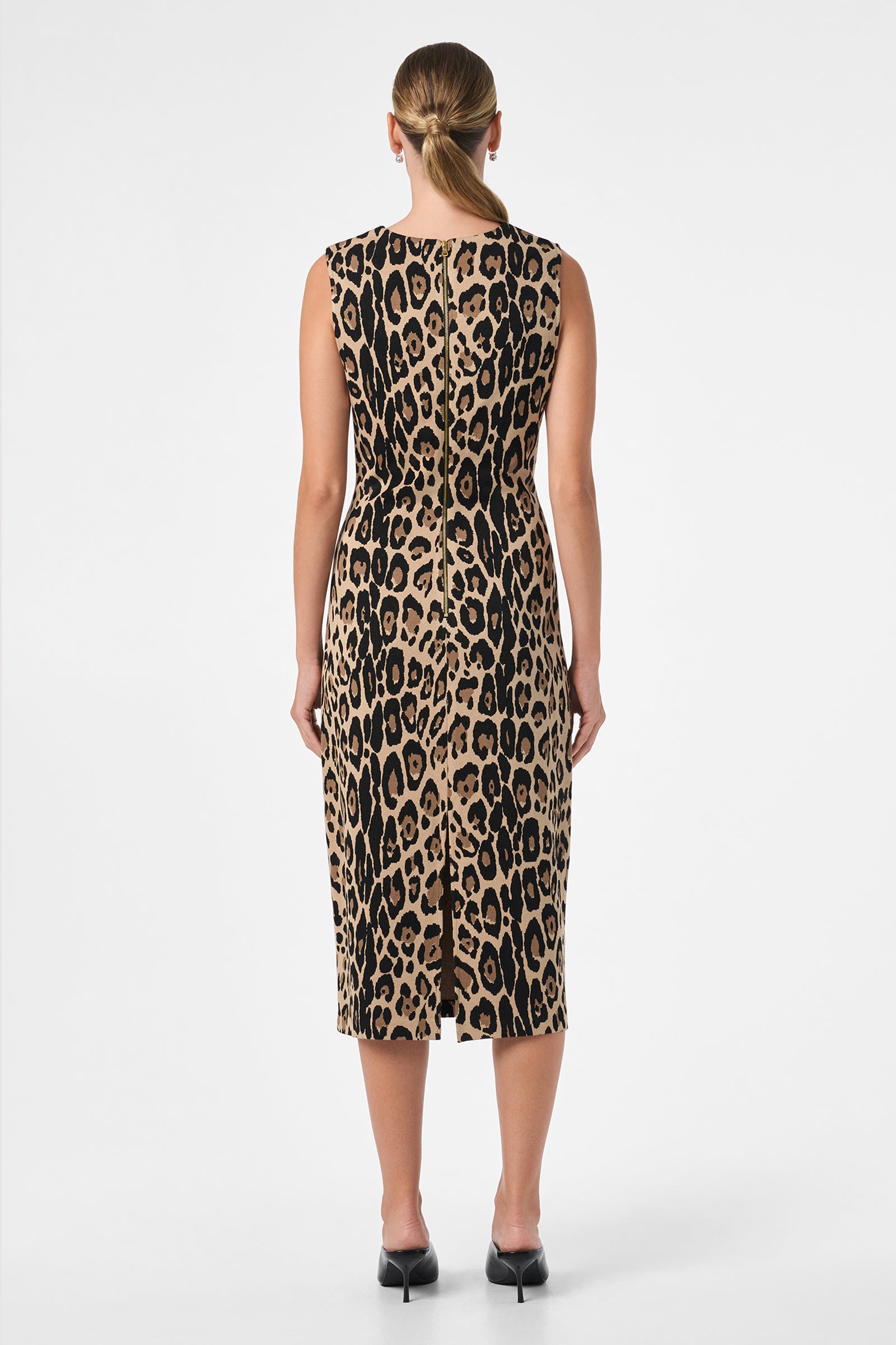 A woman with straight blonde hair in a low ponytail models the Dame Dress—a sleeveless, knee-length leopard print dress paired with black heels—standing with her back to the camera against a plain white background.
