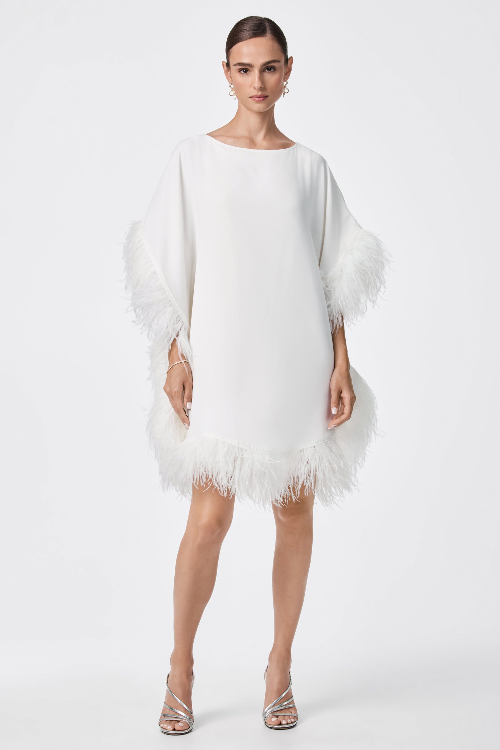 A woman wears the Mini Neena Caftan Dress, a loose white knee-length style with feather trim on the sleeves and hem. She pairs it with silver high-heeled sandals, gold hoop earrings, and a slicked-back hairstyle.