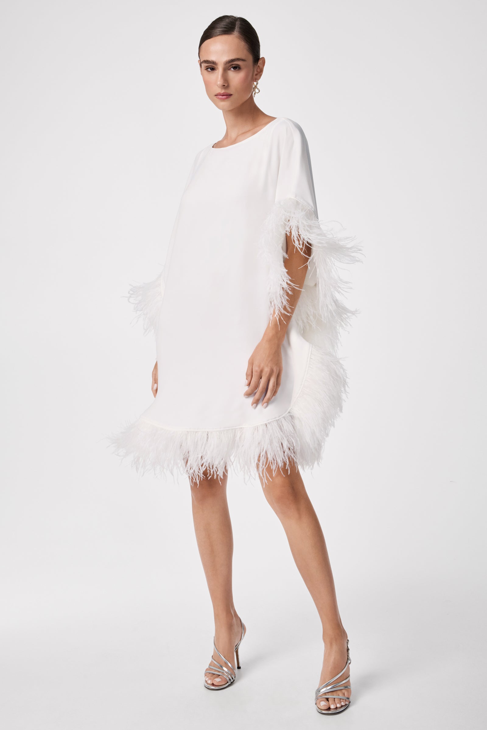 A woman stands against a plain white background wearing the Mini Neena Caftan Dress, a white, loose-fitting dress featuring feather trim on the sleeves and hem, paired with silver high-heeled sandals.