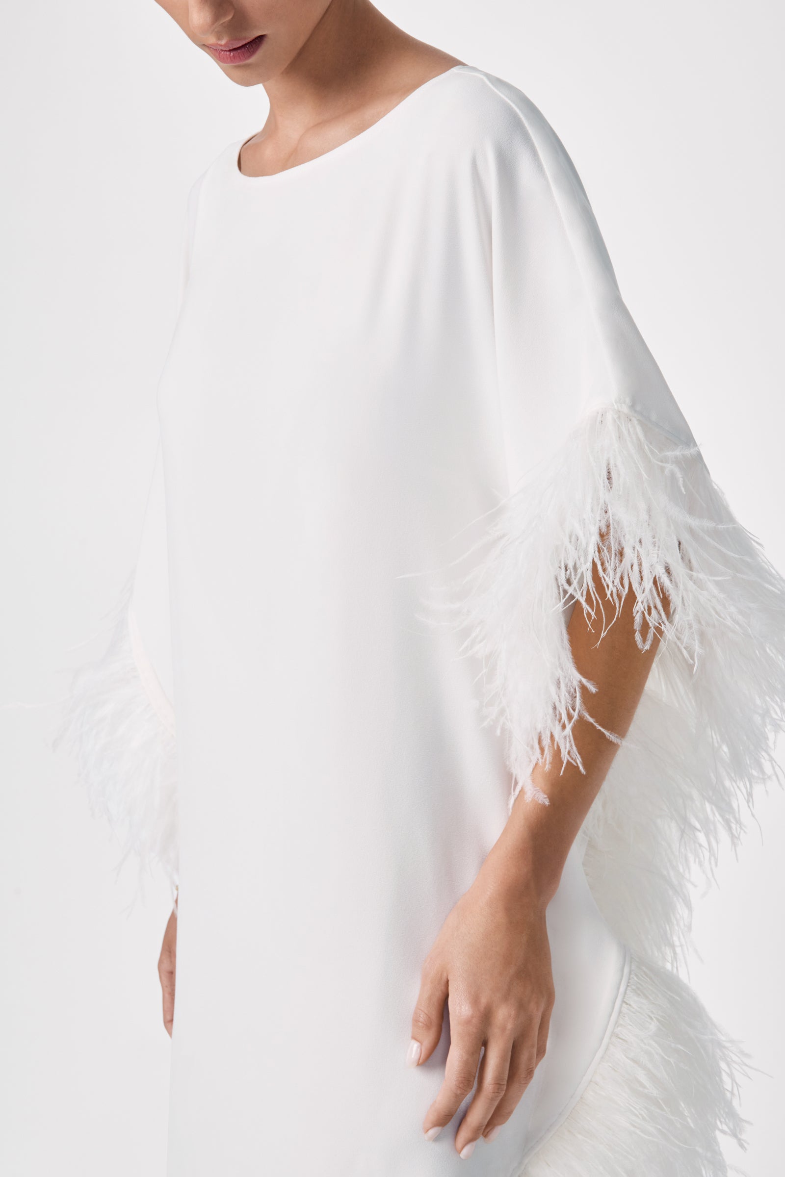 A woman wears the Mini Neena Caftan Dress—a loose, white dress with feathered sleeve trim—standing against a plain white background, her face partly out of view and her hand resting at her side.