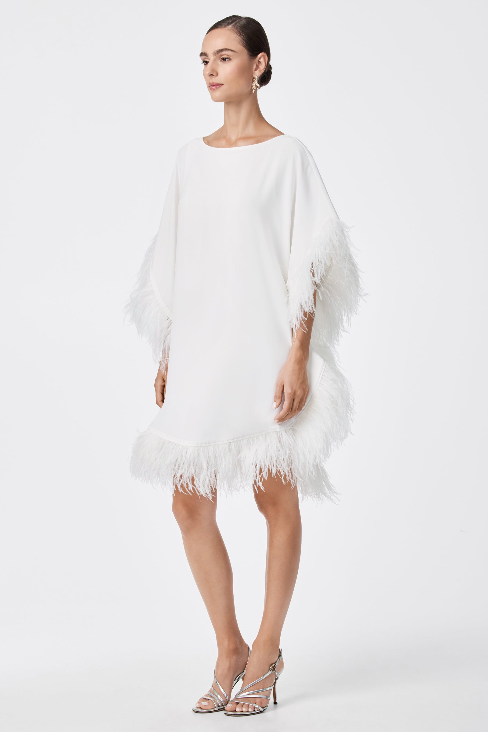 A woman wears the Mini Neena Caftan Dress, a loose white knee-length style with feather trim on the sleeves and hem, paired with silver high-heeled sandals, against a plain white background.