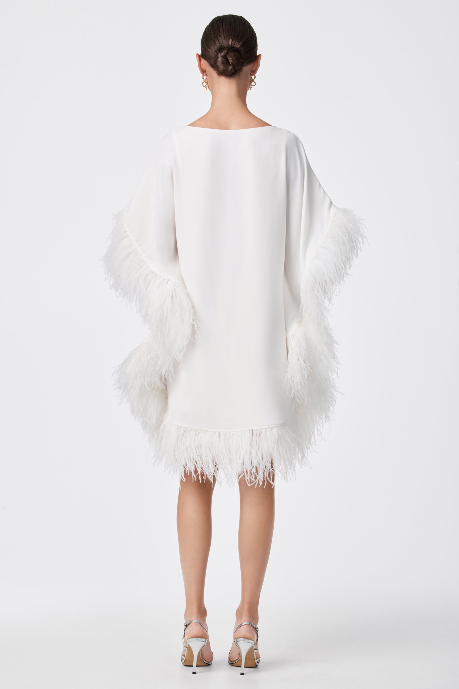 A woman faces away in the Mini Neena Caftan Dress, a loose white style with feather-trimmed sleeves and hem. Her hair is in a sleek bun, and she pairs it with silver high heels and dangling earrings against a plain white background.
