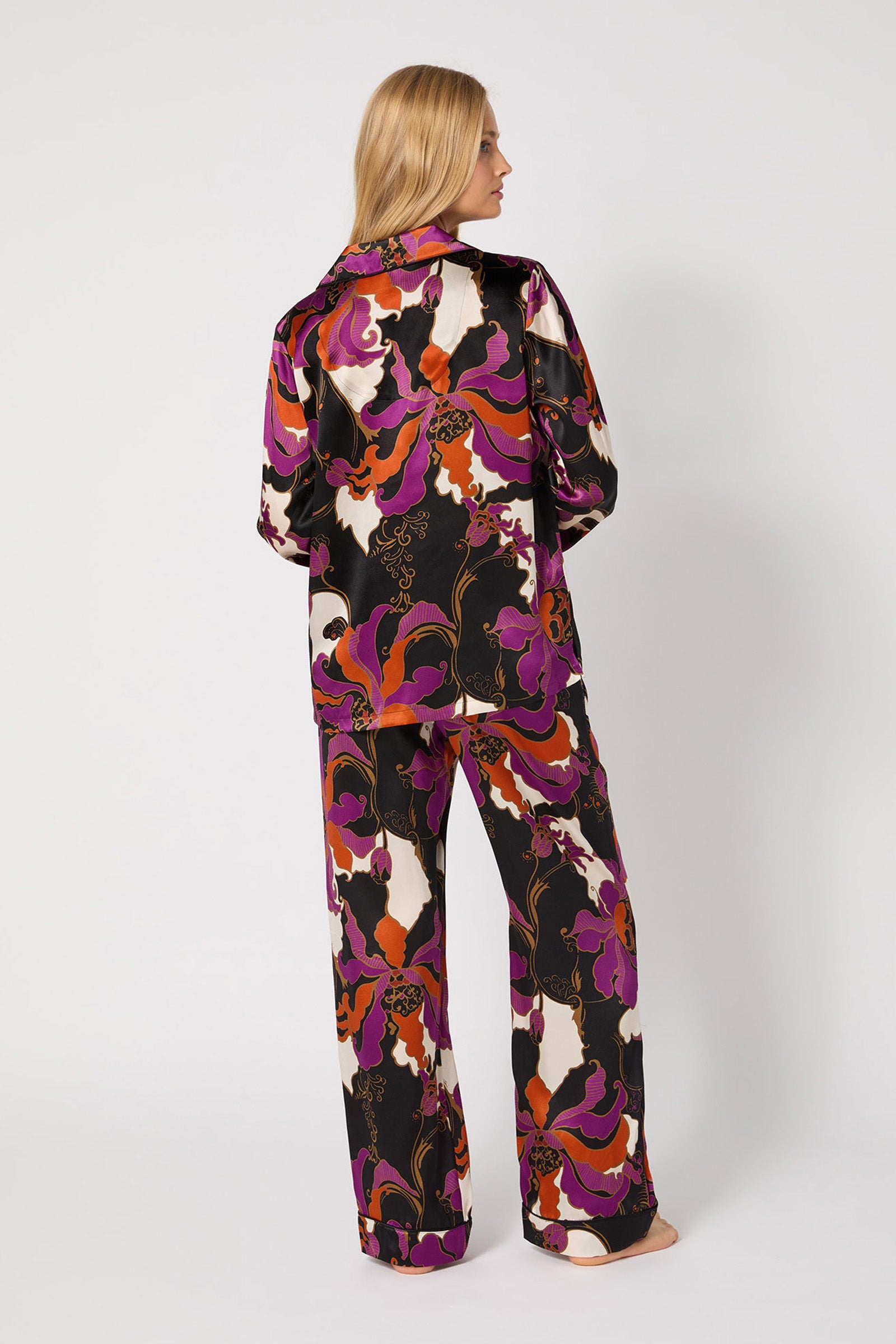 A woman stands barefoot facing away, wearing the Classic Silk PJ Set - Fall Floral: loose-fitting matching satin pajamas with a bold purple, orange, white, and black floral print. The background is plain white.