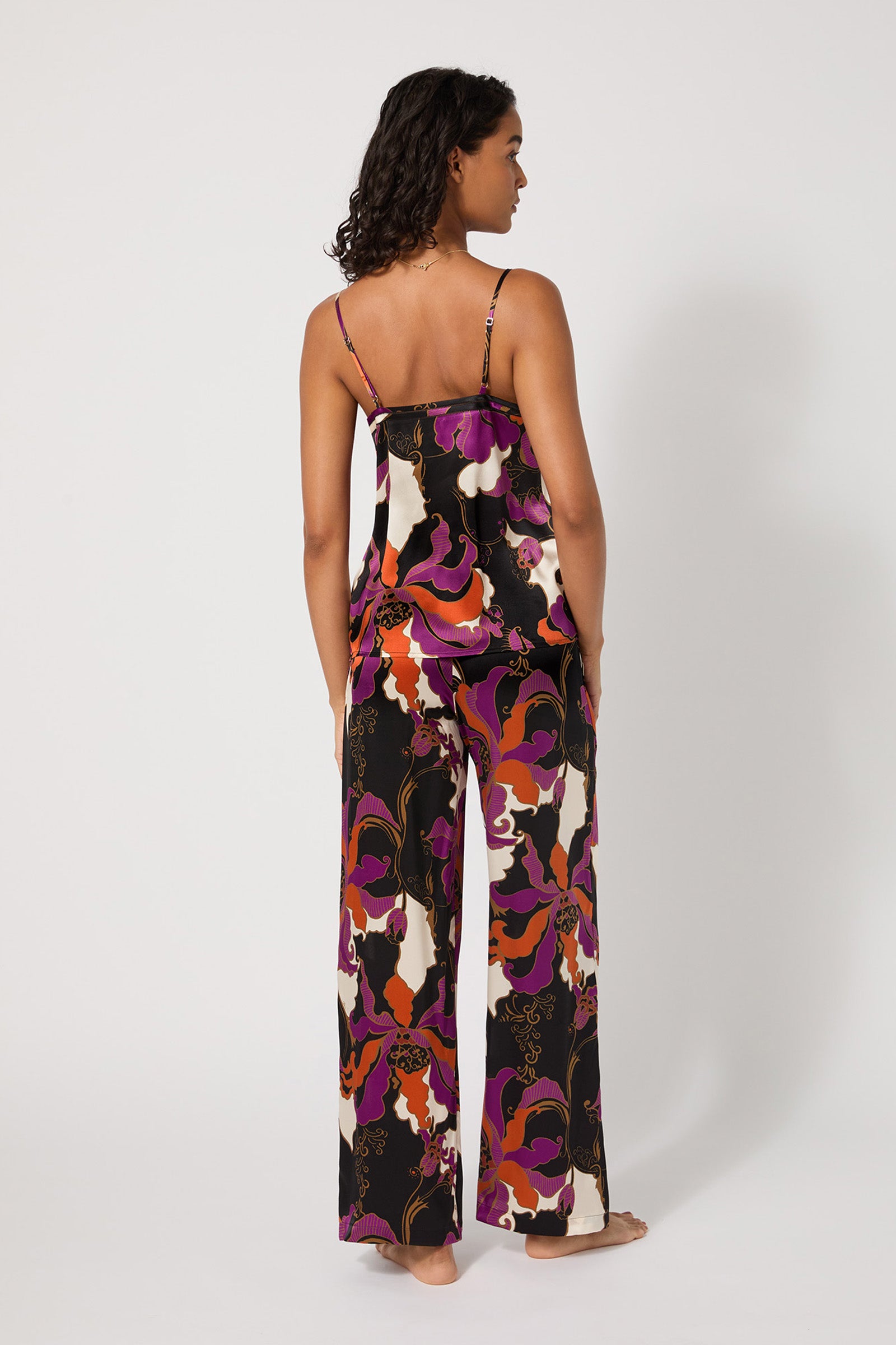 A woman stands barefoot, facing away, wearing the Silk Cami PJ Set - Fall Floral featuring a colorful abstract floral pattern in black, purple, orange, and white. The background is plain white.