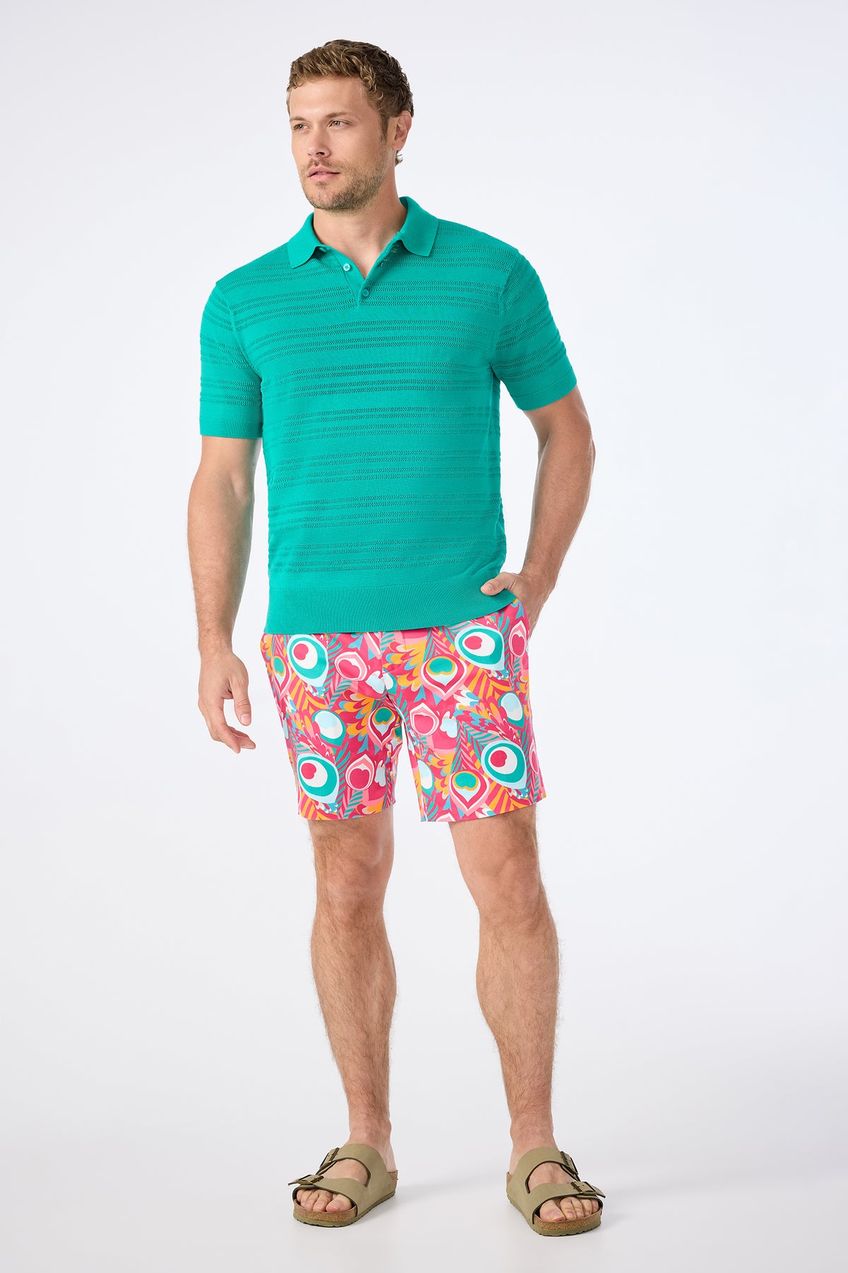 A man wears Dramont Herringbone Lawrence Shorts, a teal short-sleeve knit polo, and tan sandals while standing against a plain white background.