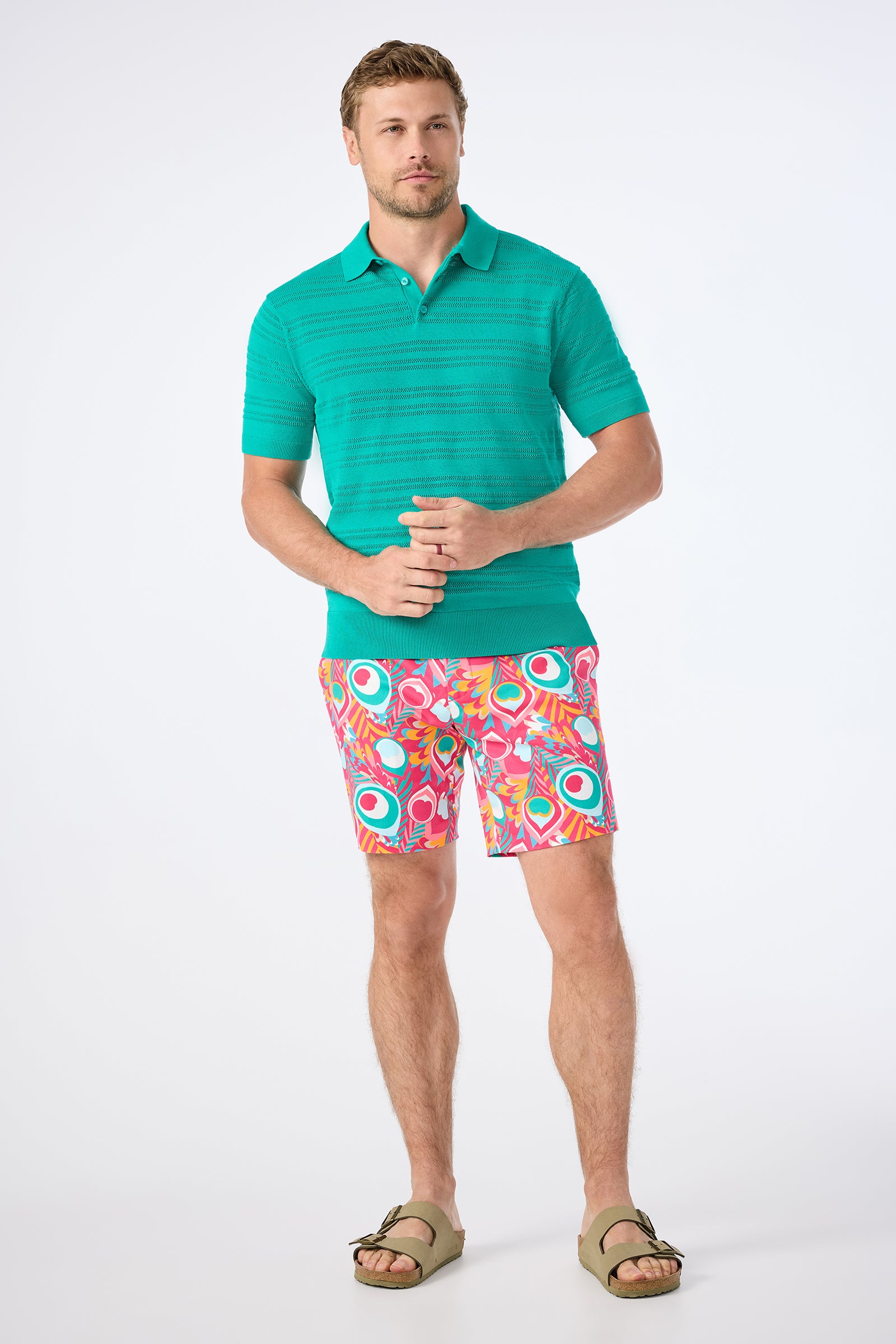 A man with short light brown hair and a relaxed expression stands against a white background wearing the Edison Short-Sleeve Striped Polo, bright patterned shorts with circular designs, and olive green sandals.