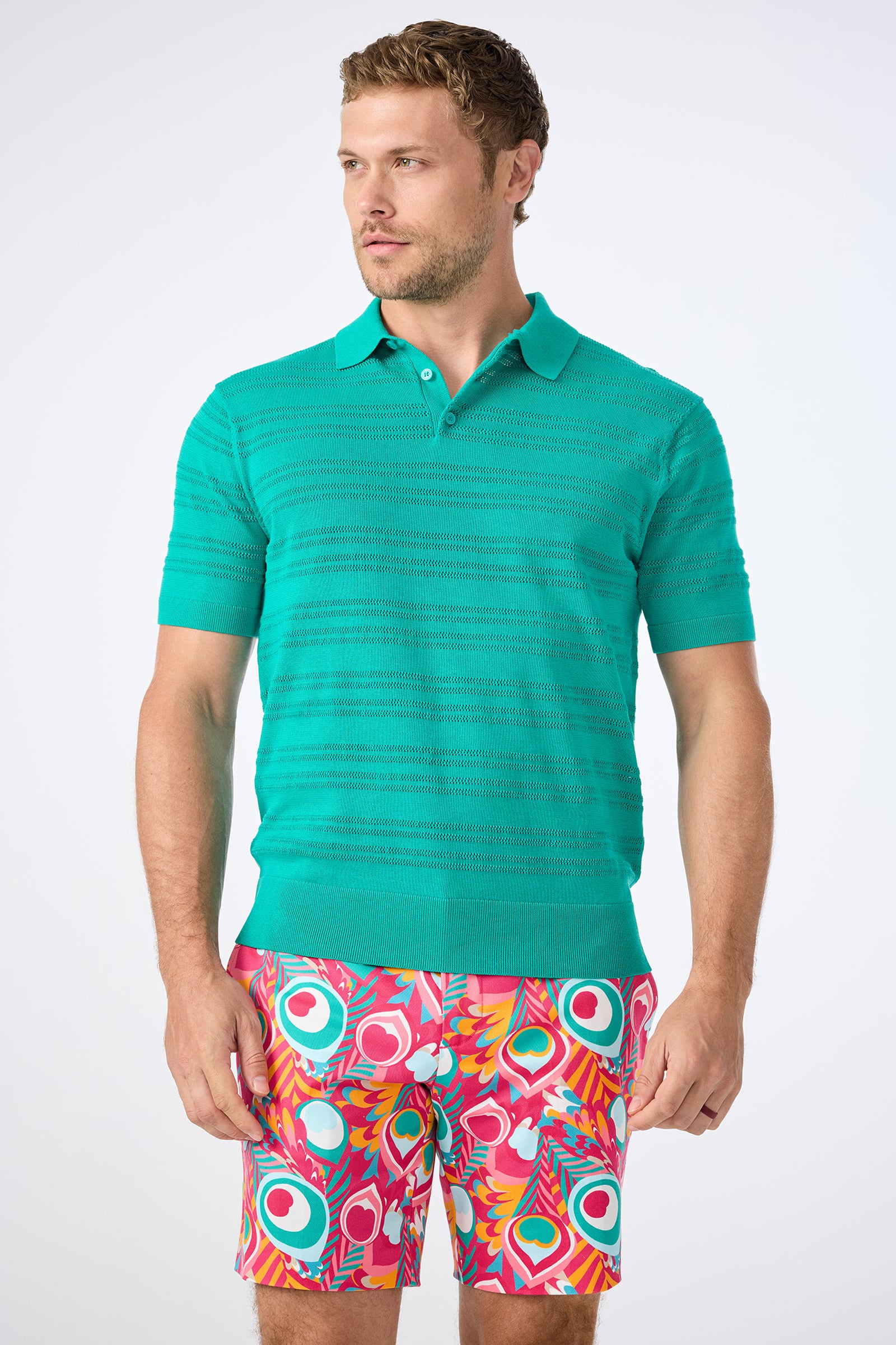 A man wears the Edison Short-Sleeve Striped Polo and colorful abstract-patterned shorts while standing against a plain white background.