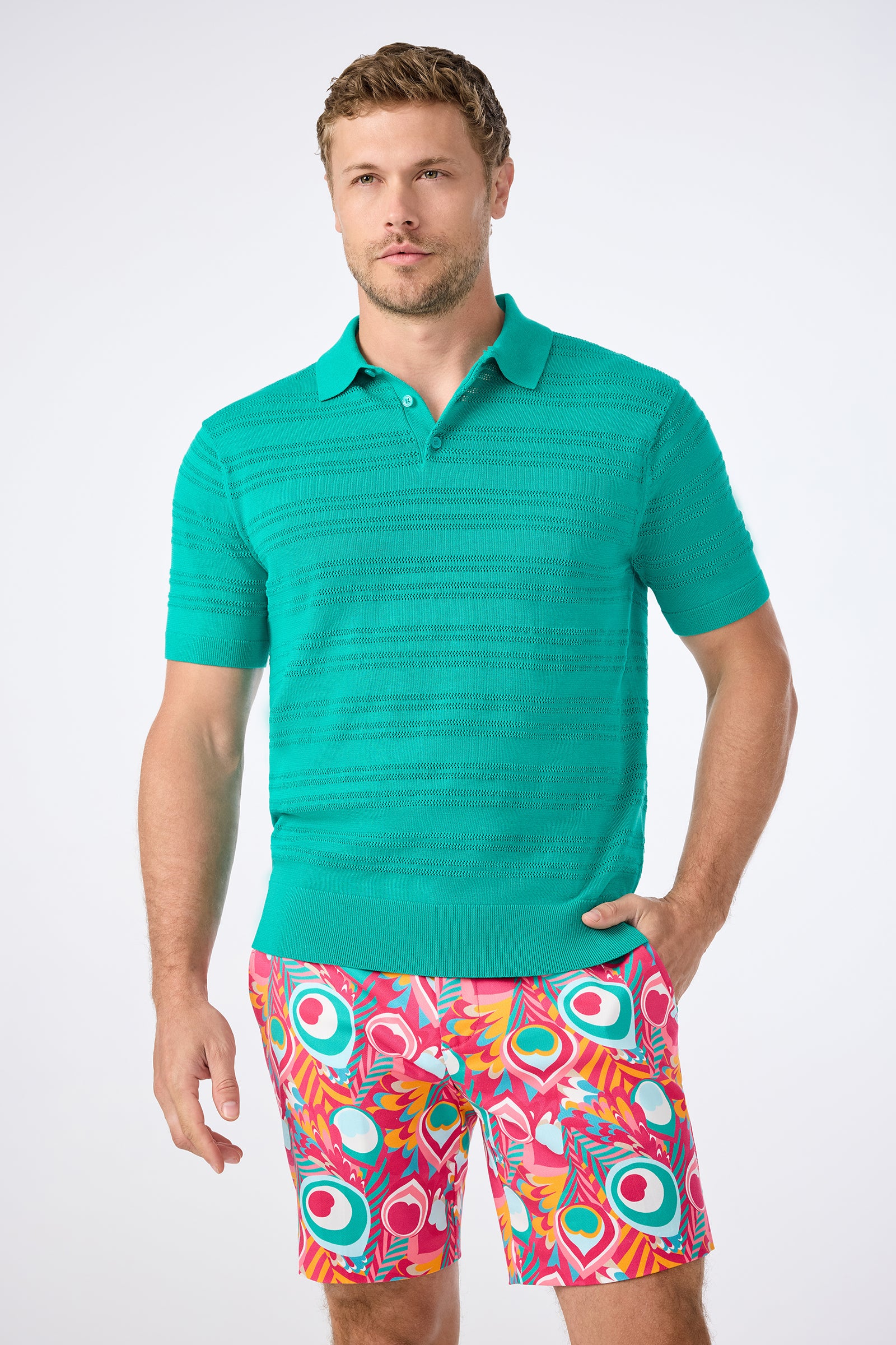 A man wears the Edison Short-Sleeve Striped Polo in Toulon Teal and bold, colorful patterned shorts, standing against a plain light background with one hand in his pocket.