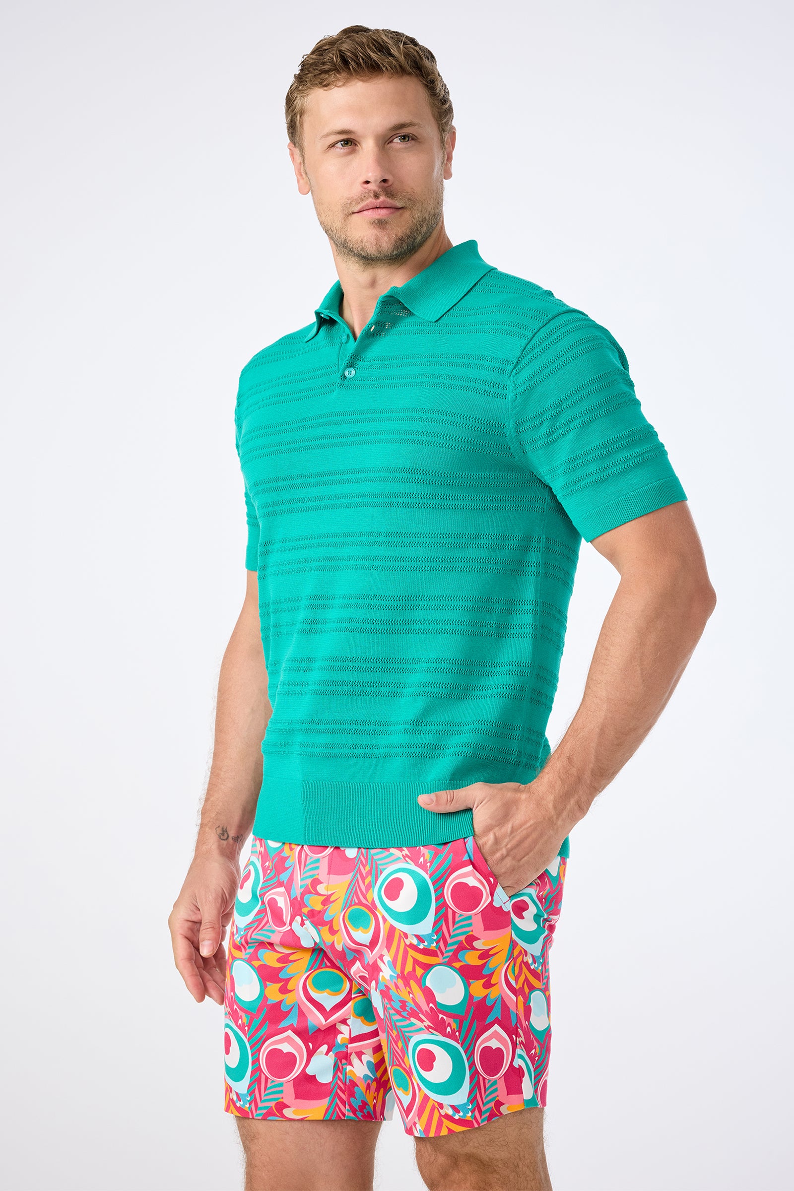 A man wearing an Edison Short-Sleeve Striped Polo and colorful patterned shorts stands with one hand in his pocket, looking slightly off camera against a plain light background.
