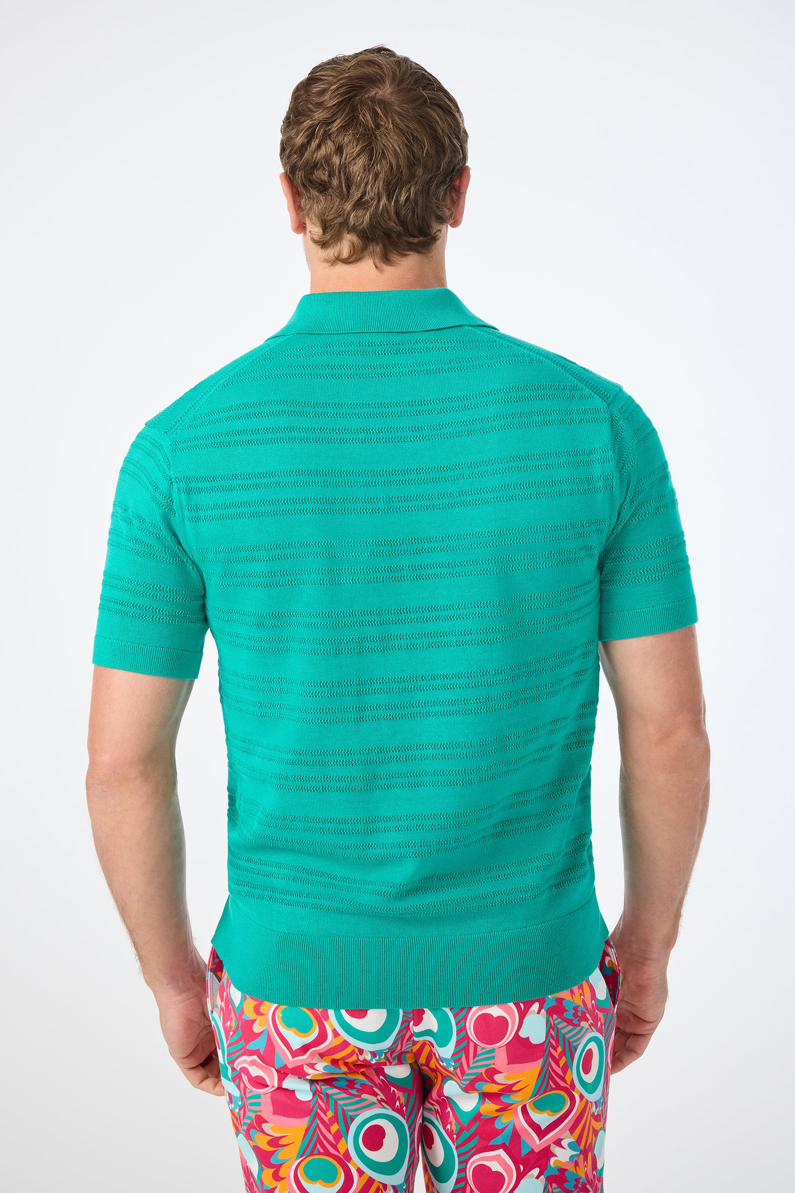 A person with short, light brown hair is seen from behind, wearing the Edison Short-Sleeve Striped Polo and colorful abstract-patterned pants featuring red, yellow, white, and teal shapes on a white background.