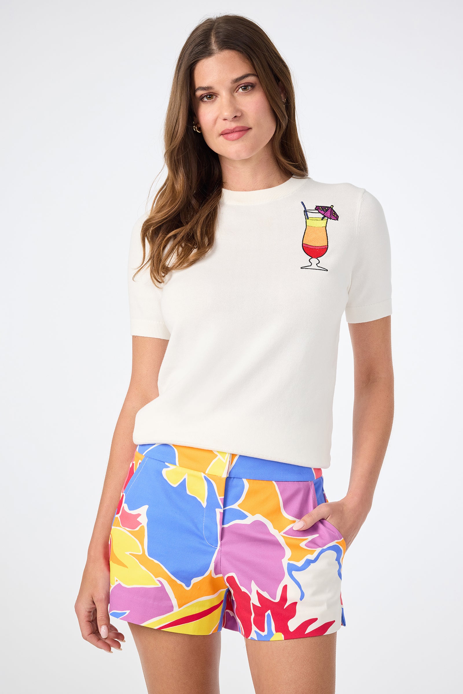 A woman wears the Old Fashioned Crewneck in Whitewash, featuring a colorful cocktail graphic on the chest, paired with bright multicolored floral shorts, standing against a plain light background.