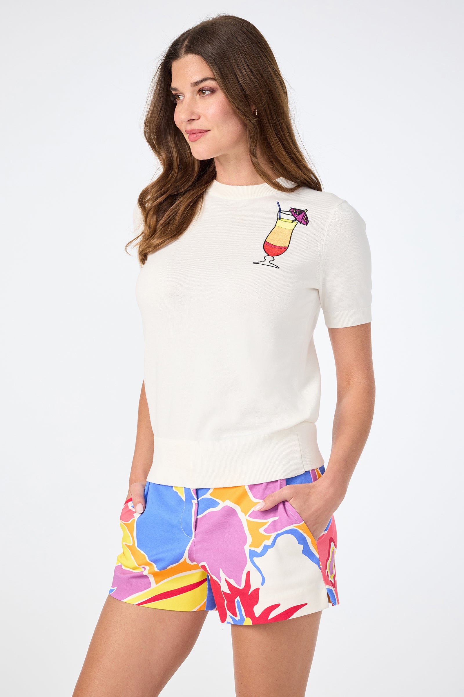 A woman with long brown hair wears the Old Fashioned Crewneck, a white short-sleeve sweater featuring a colorful cocktail graphic, paired with bright abstract multicolored shorts. She stands hands-in-pockets, gazing to the side.
