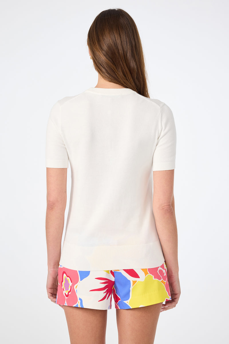 A woman with long brown hair, seen from behind, wears the Old Fashioned Crewneck white short-sleeve top and colorful floral print shorts featuring red, yellow, blue, and pink flowers on a white background.