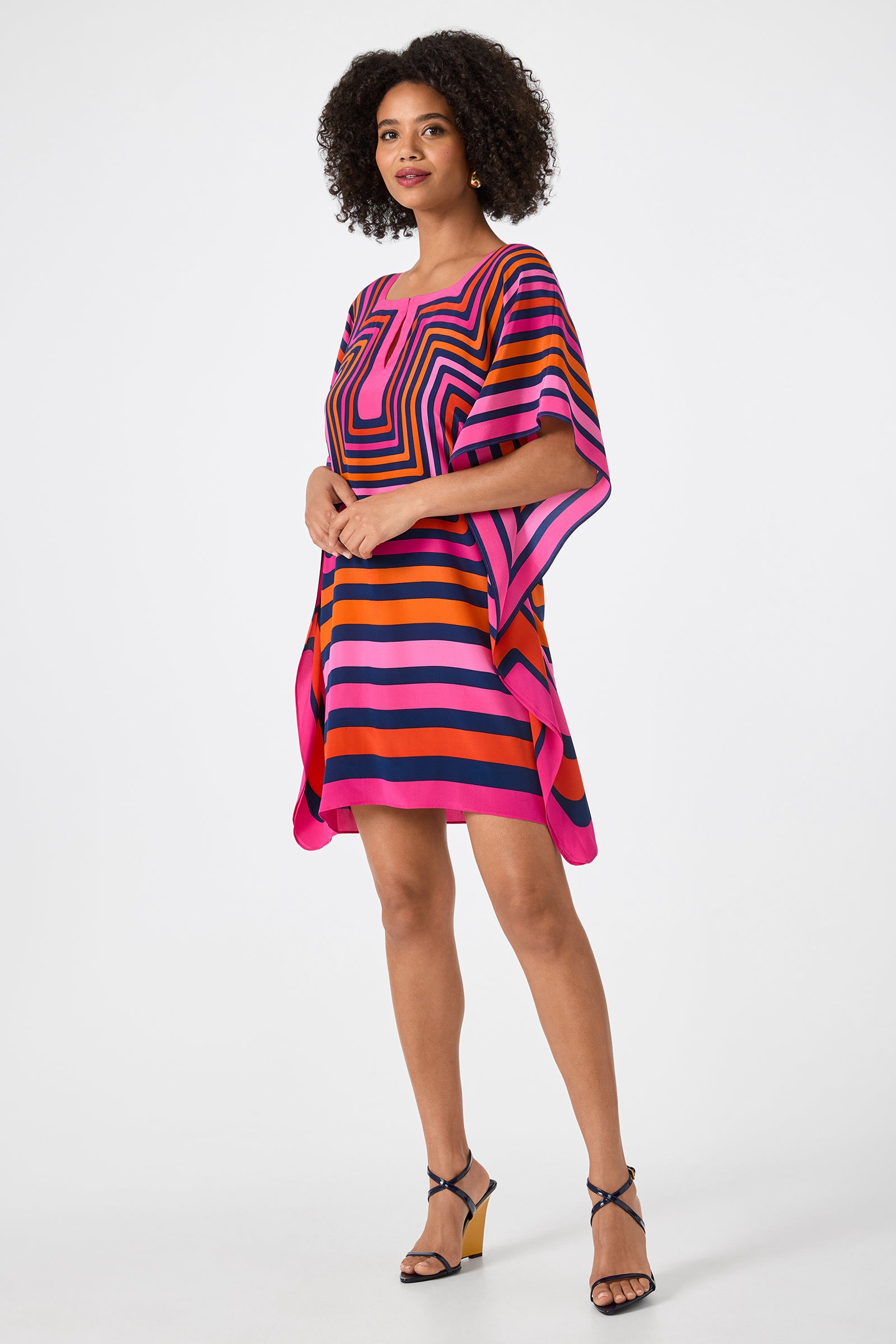 A woman stands against a plain white background wearing the Theodora Caftan Dress - Gaki Geo, featuring colorful geometric patterns in pink, orange, black, and blue stripes, paired with strappy wedge sandals.