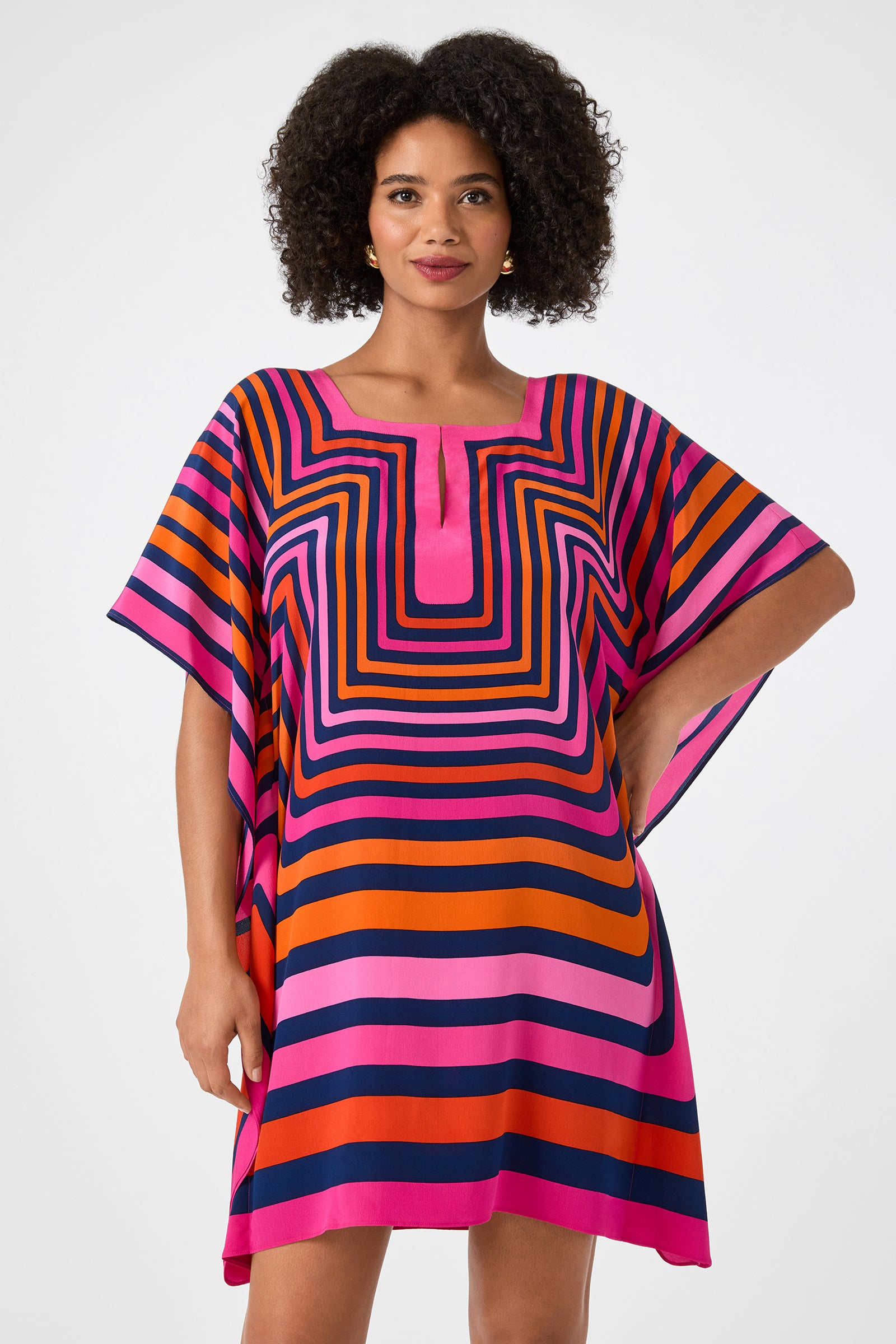 A woman stands with one hand on her hip, wearing the Theodora Caftan Dress - Gaki Geo. This loose, short dress features bold pink, orange, purple, and navy geometric stripes, wide sleeves, a square neckline, and is set against a plain white background.