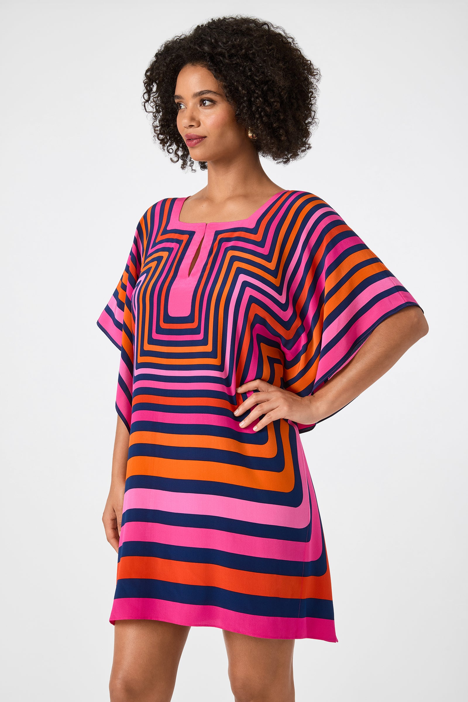 A woman with curly hair wears the Theodora Caftan Dress - Gaki Geo, featuring colorful pink, orange, navy blue, and black geometric stripes. She stands with one hand on her hip against a plain background.