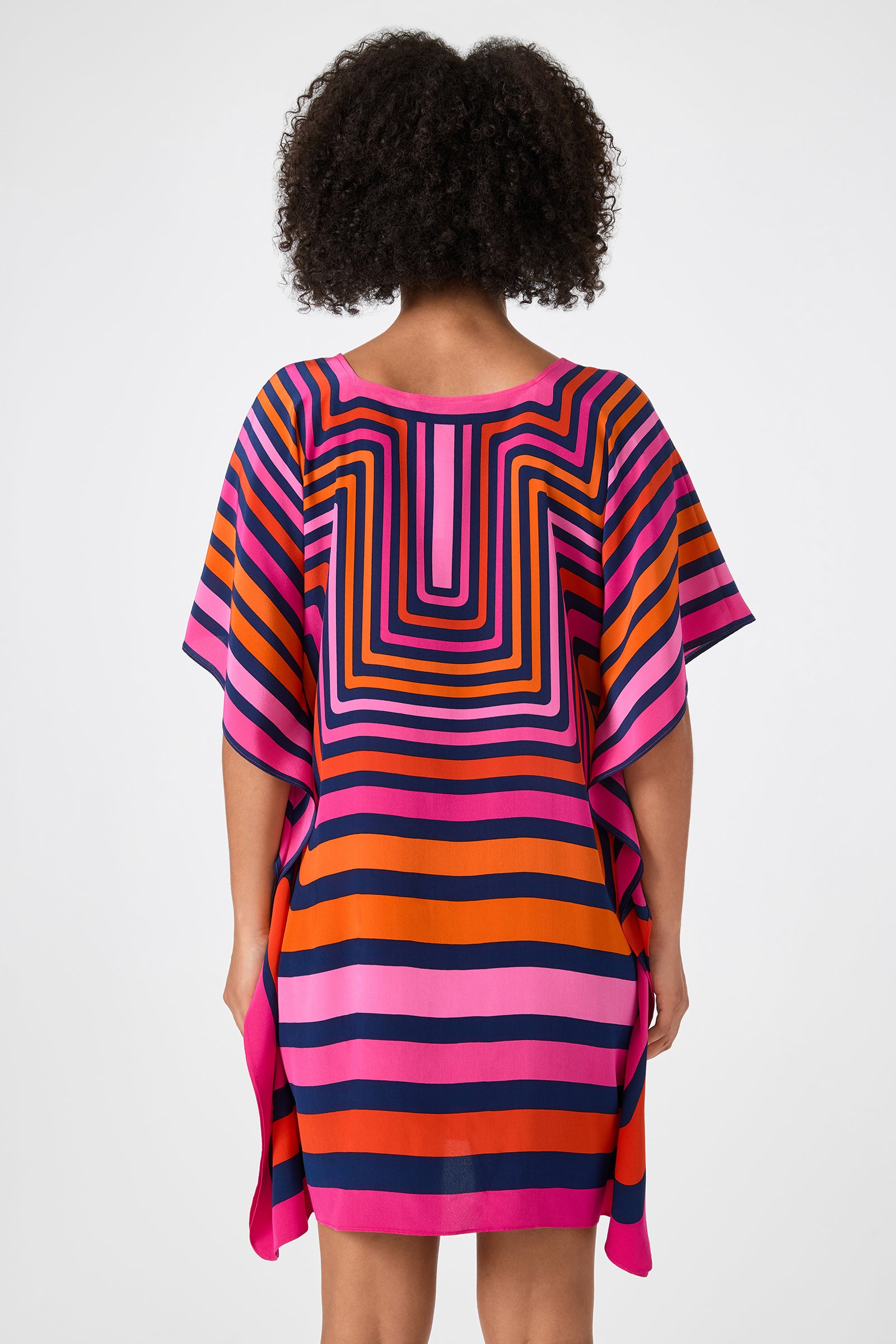 A person with curly hair is shown from the back, wearing Theodora Caftan Dress - Gaki Geo, a loose, short-sleeved dress featuring bold pink, orange, navy, and purple geometric stripes on a white background.
