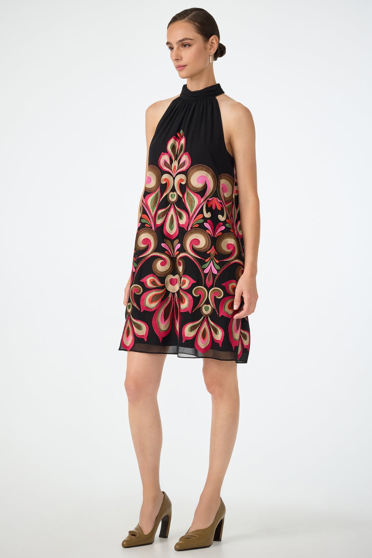 A woman wears the Zana Embroidered Mini Dress, featuring a black halter-neck design with ornate pink, red, and gold floral embroidery, paired with tan high heels against a plain white background.