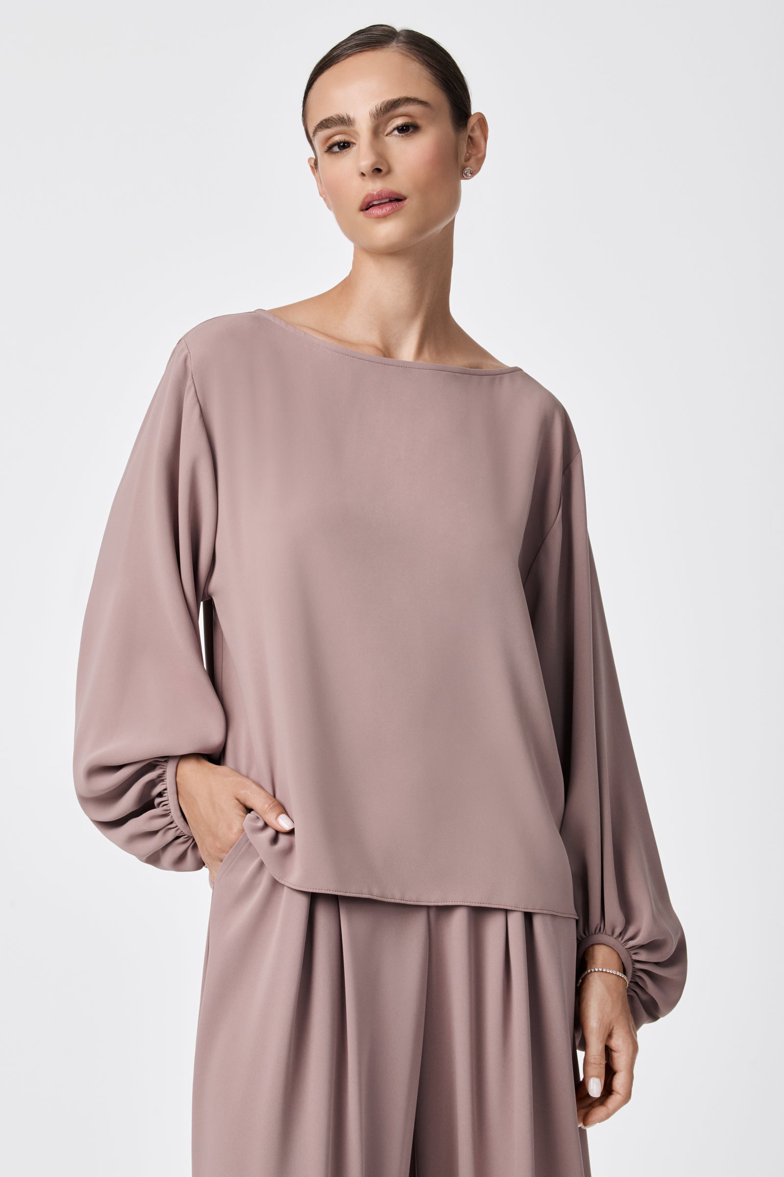 A woman models the Ursa 2 Balloon Sleeve Top—a loose, mauve blouse with billowy long sleeves—paired with matching wide-leg pants, one hand in her pocket, standing against a plain, light background.