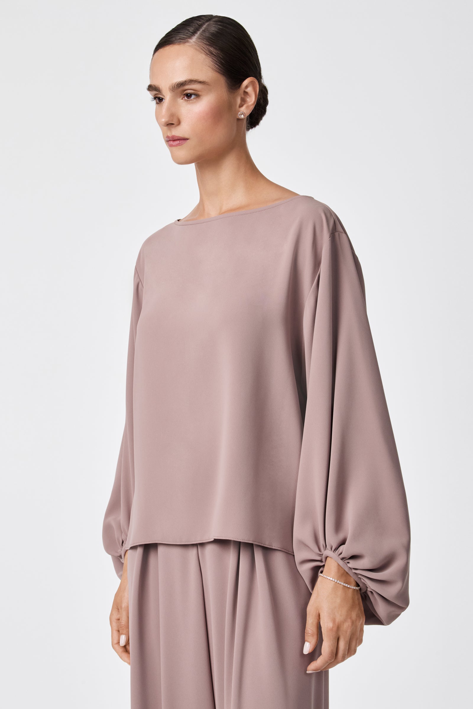 A woman with her hair in a neat bun stands against a white background, wearing the URSA 2 TOP—a loose-fitting, long-sleeved taupe blouse made from 100% polyester—paired with matching pants.