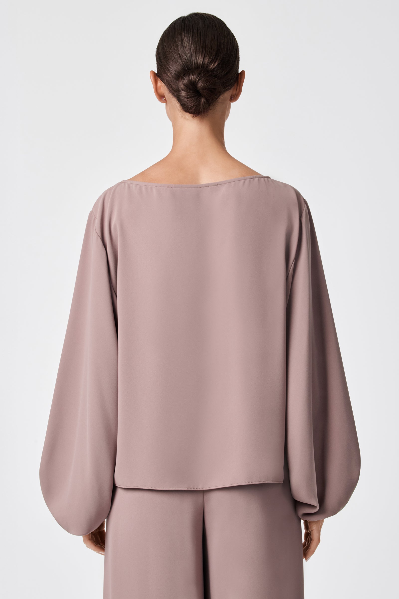 A woman with dark hair in a low bun is seen from behind, wearing the URSA 2 TOP—a loose, long-sleeved blouse in mauve, crafted from soft polyester with a minimalist design and paired with matching pants.