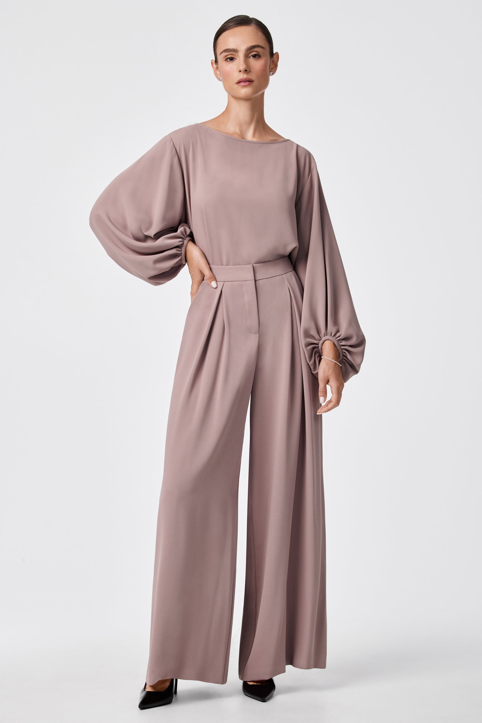 A woman stands against a plain background wearing the URSA 2 TOP, a mauve long-sleeve blouse with balloon sleeves, paired with matching wide-leg pants and black pointed-toe heels.