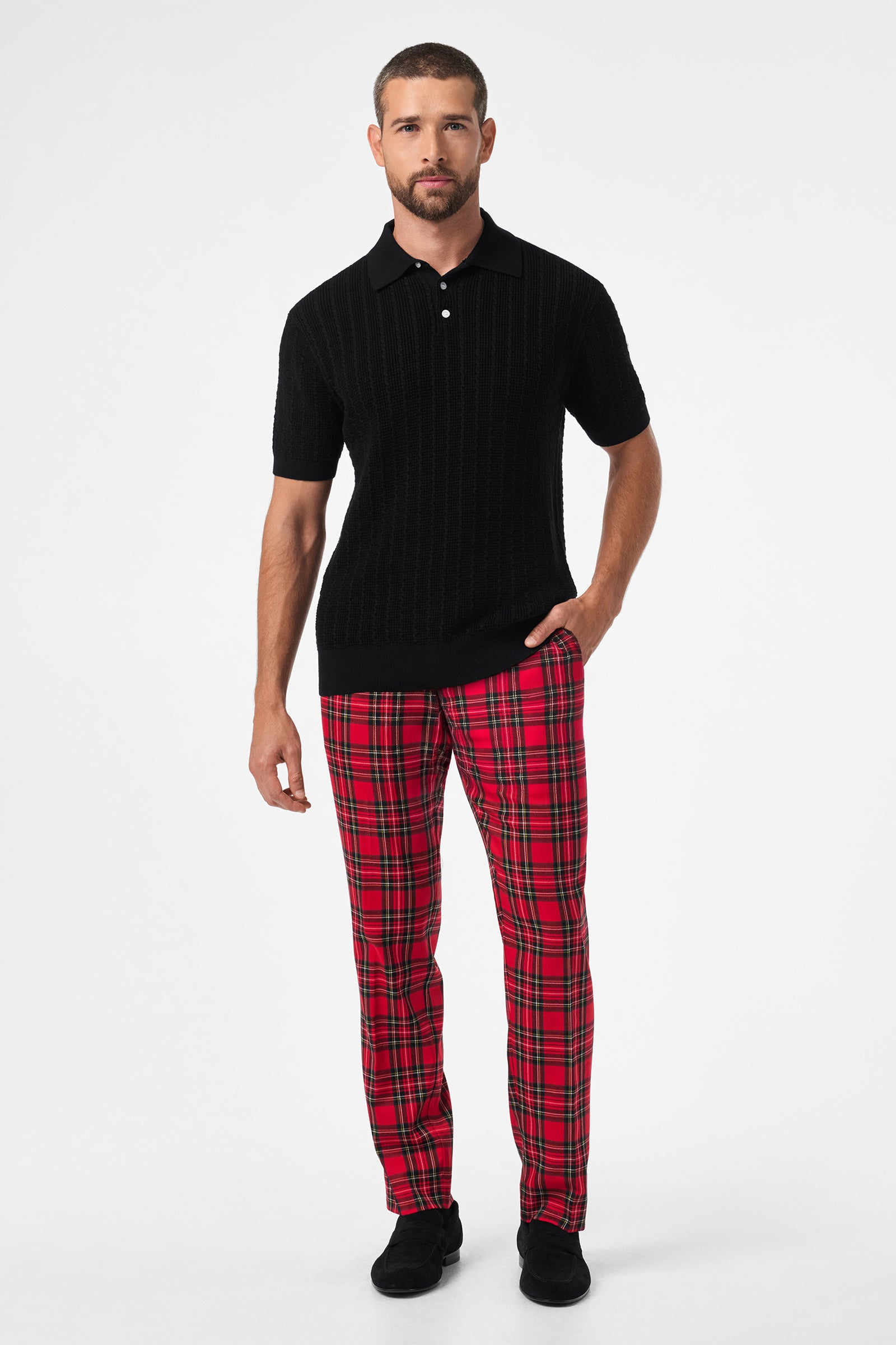 A man with short hair and a beard stands against a white background, wearing a black short-sleeve knit polo, black shoes, and the Clyde Slim Trouser - Can-Can Check. One hand is in his pocket; his expression is neutral.