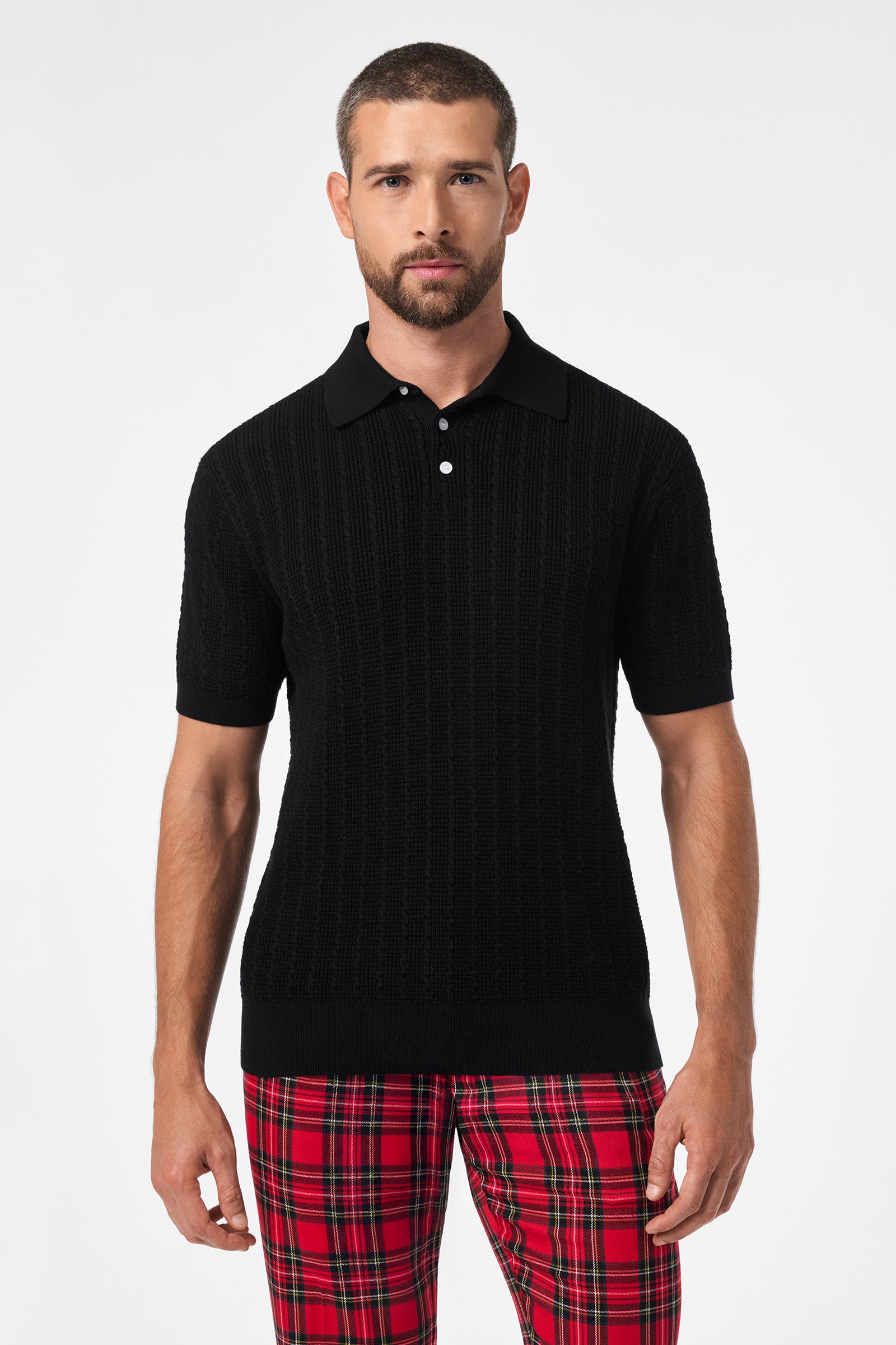 A man with short hair and a beard wears the black Asher Short-Sleeve Polo and red plaid pants, standing against a plain white background.