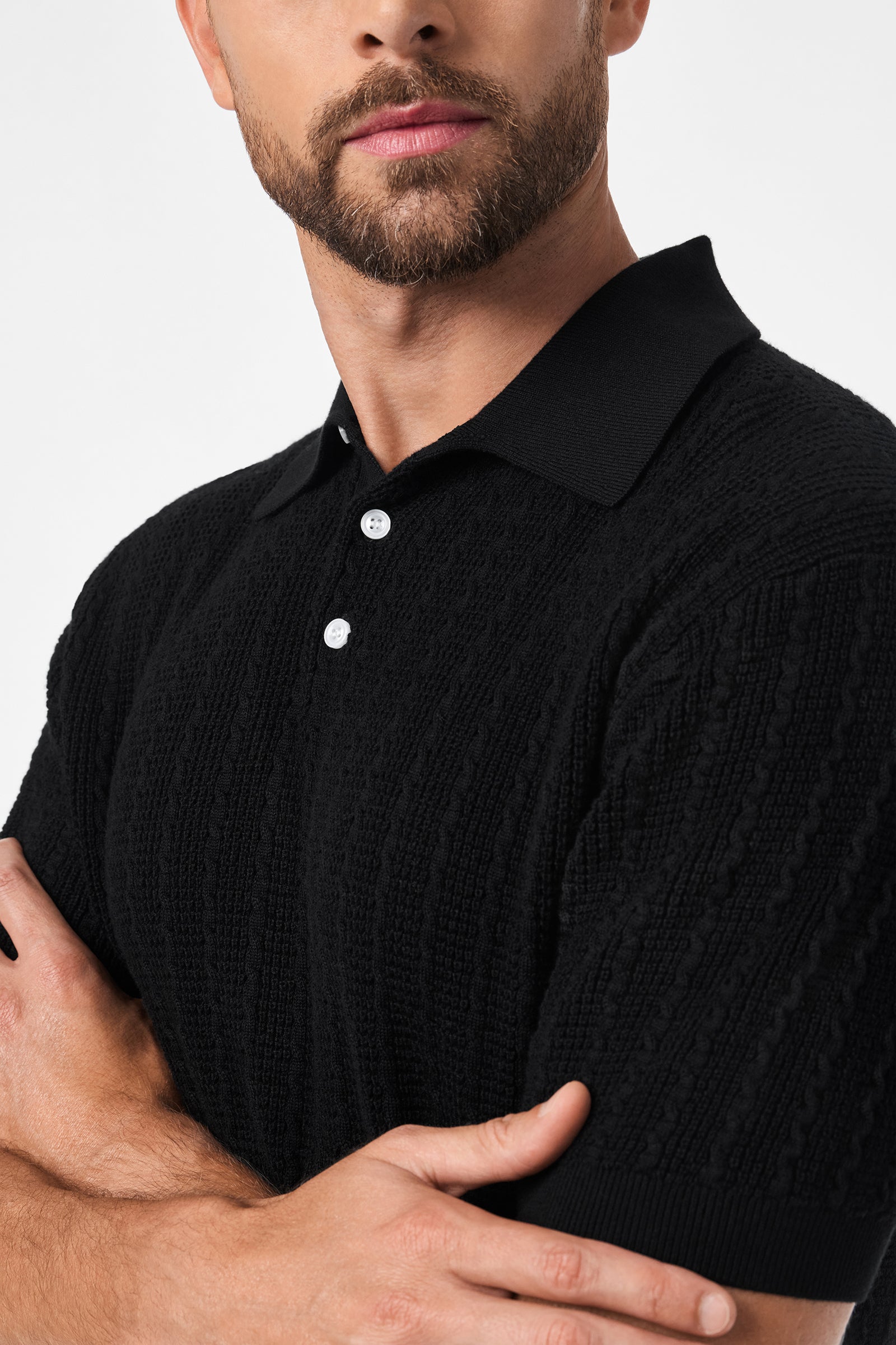 A man with a trimmed beard and mustache stands with arms crossed against a plain white background, wearing the black Asher Short-Sleeve Polo featuring a textured fabric, collar, and white buttons.