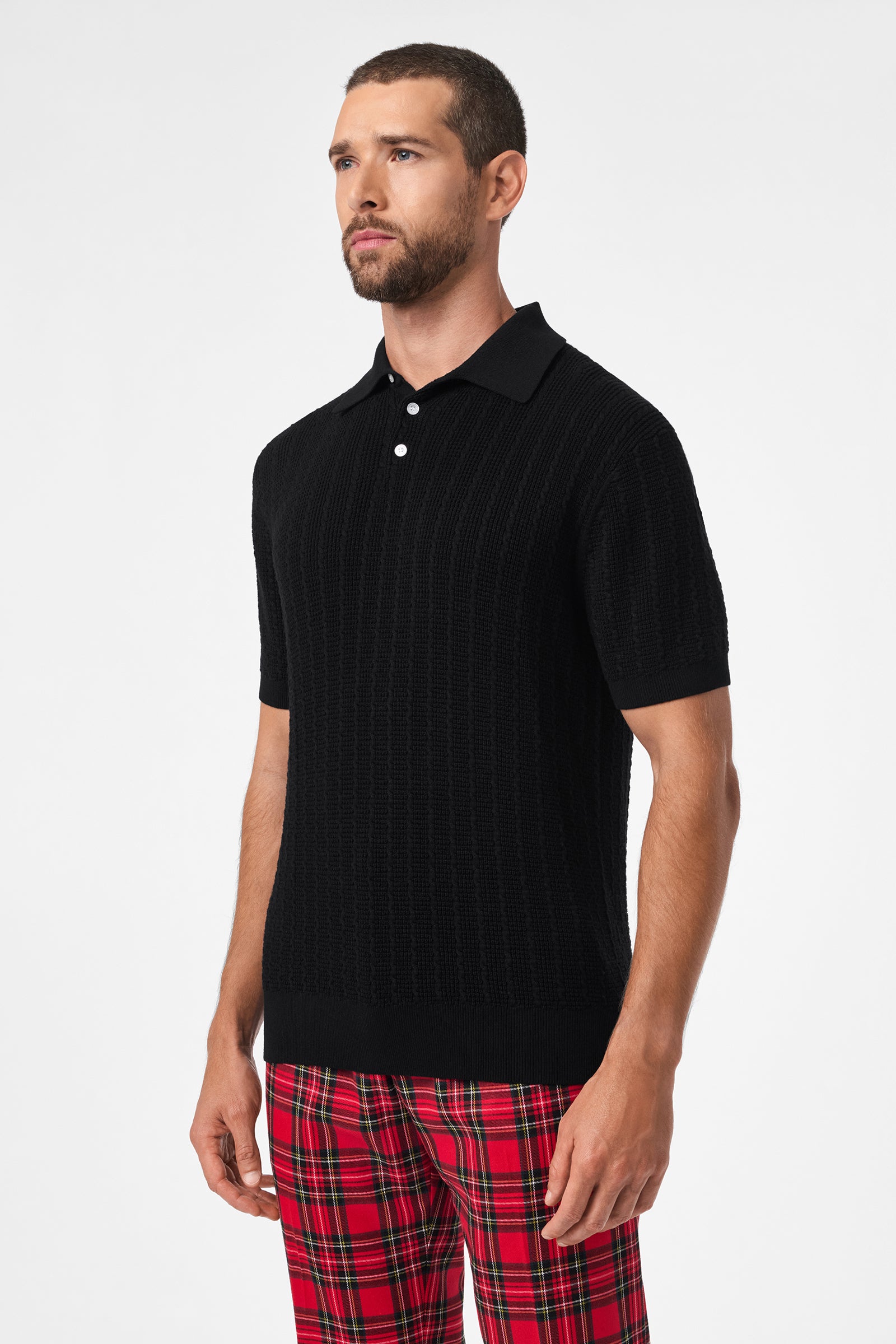 A man with short hair and a beard wears the Asher Short-Sleeve Polo in black and red plaid pants, standing against a plain white background.
