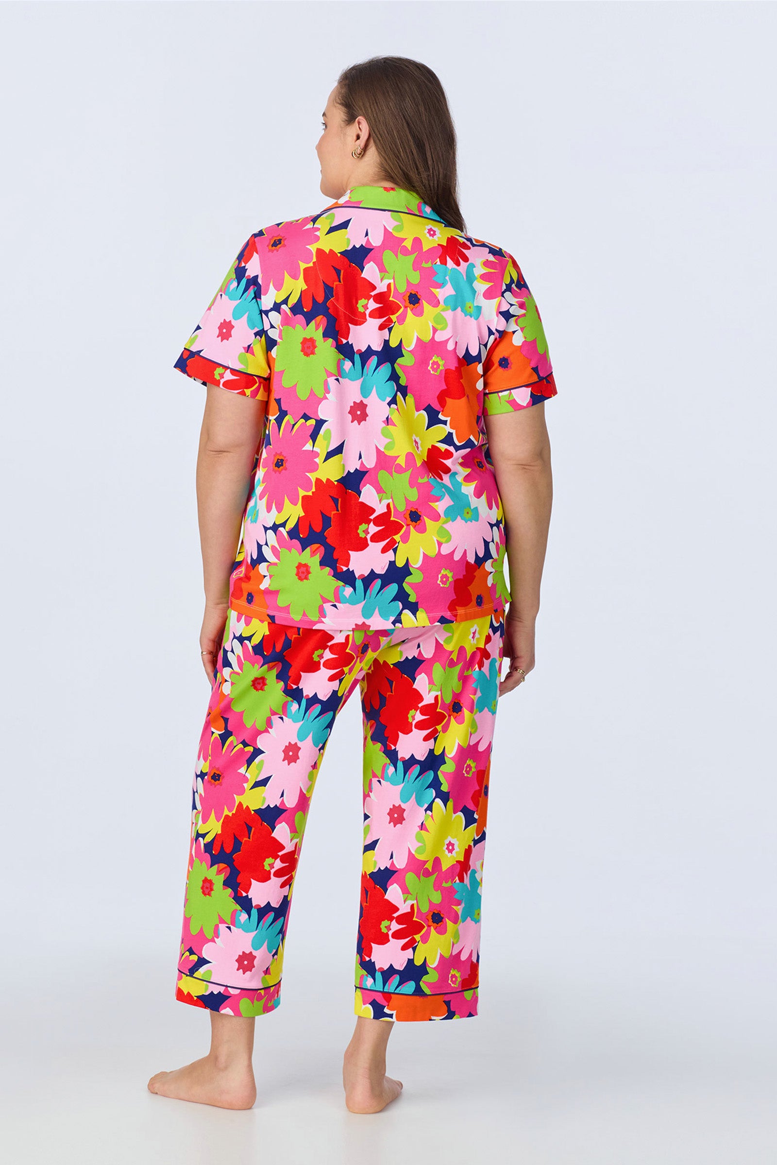 A barefoot person faces away, wearing the Crop Classic PJ Set - Pop Floral Stretch Jersey featuring a colorful floral pattern in red, pink, green, yellow, blue, and white on both the short-sleeve top and pants.